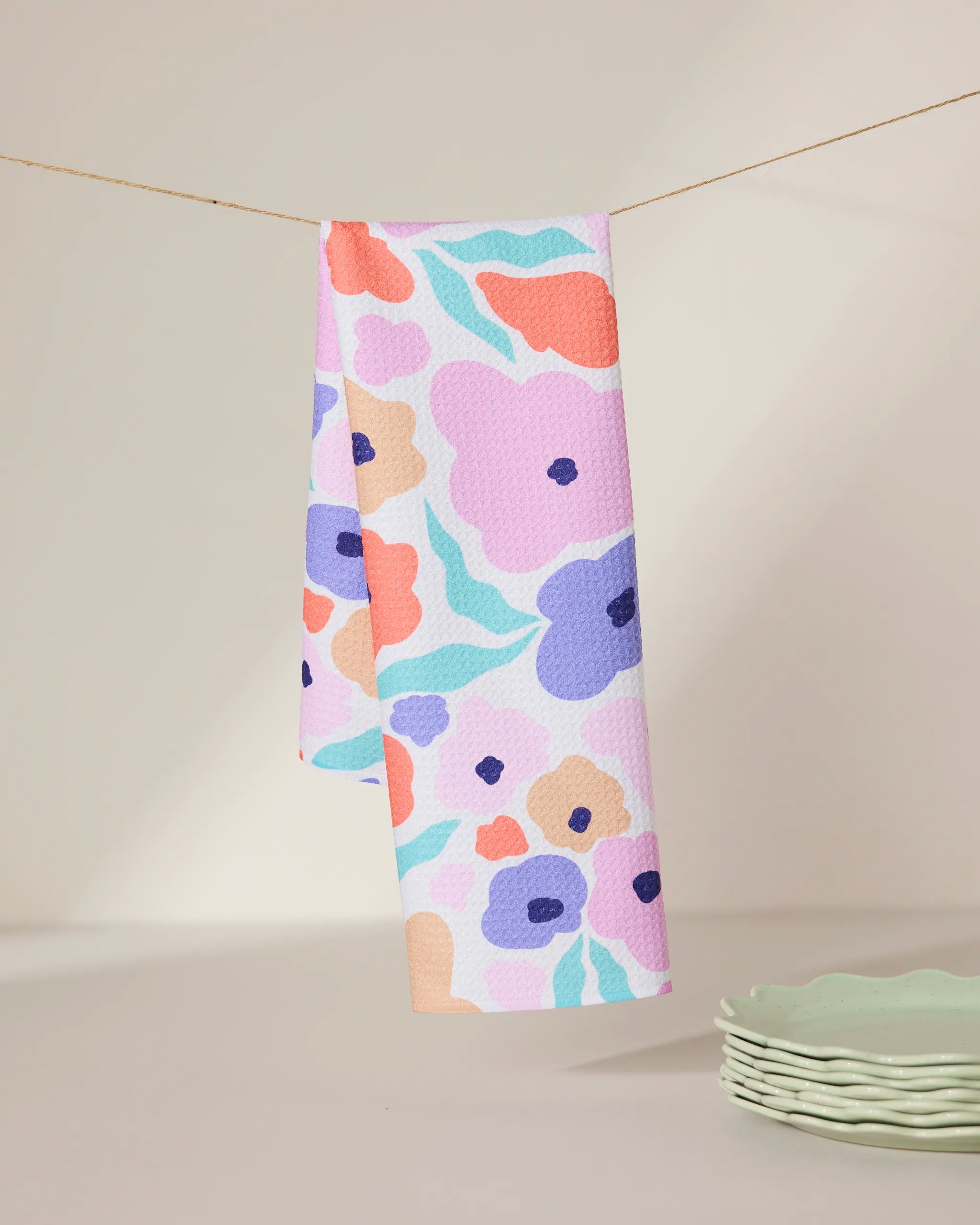 The GEOMETRY Blossoming Love tea towel, made from recycled materials and featuring a colorful floral pattern, hangs on a string against a neutral background with light green scalloped plates stacked nearby.