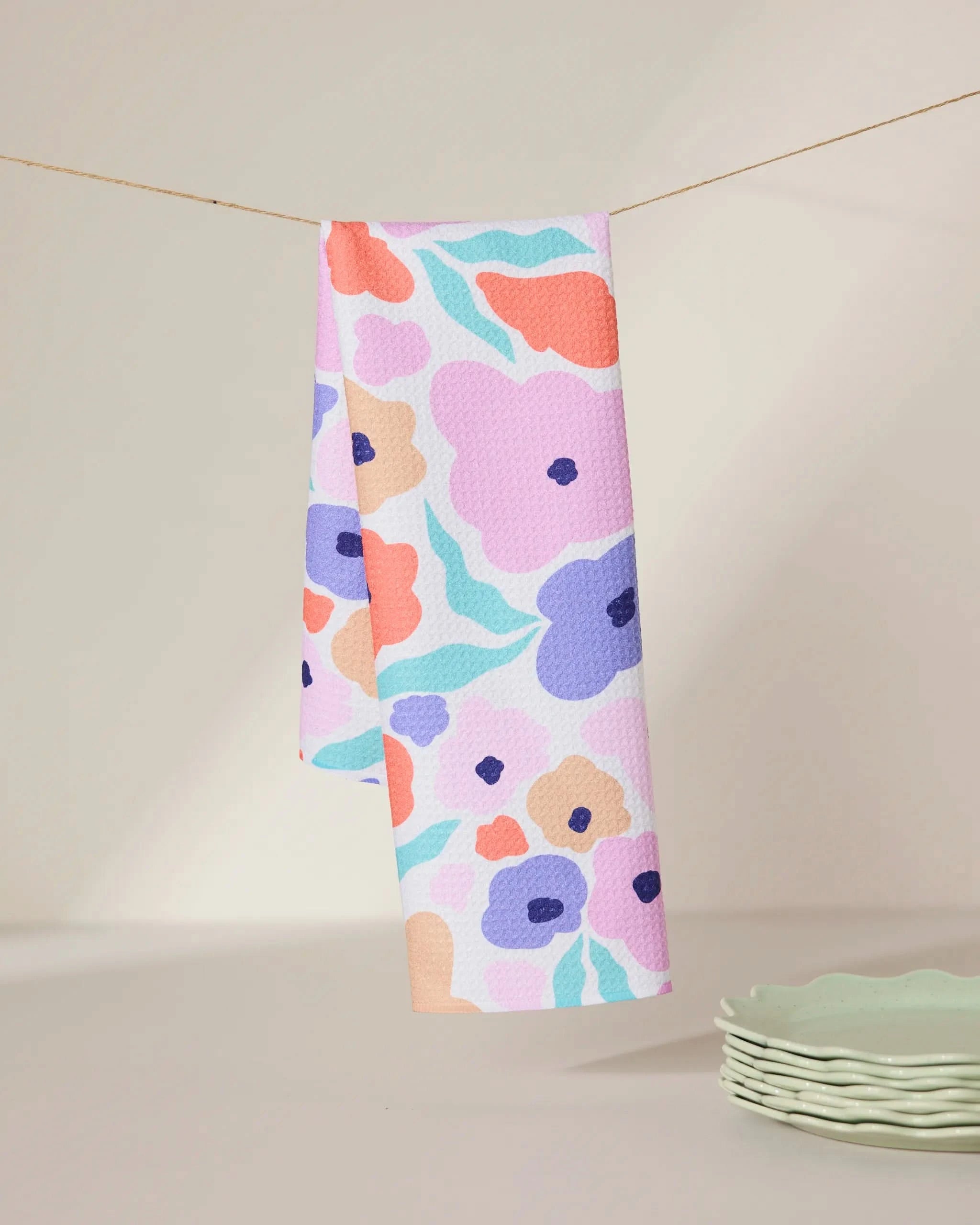 The GEOMETRY Blossoming Love tea towel, made from recycled materials and featuring a colorful floral pattern, hangs on a string against a neutral background with light green scalloped plates stacked nearby.