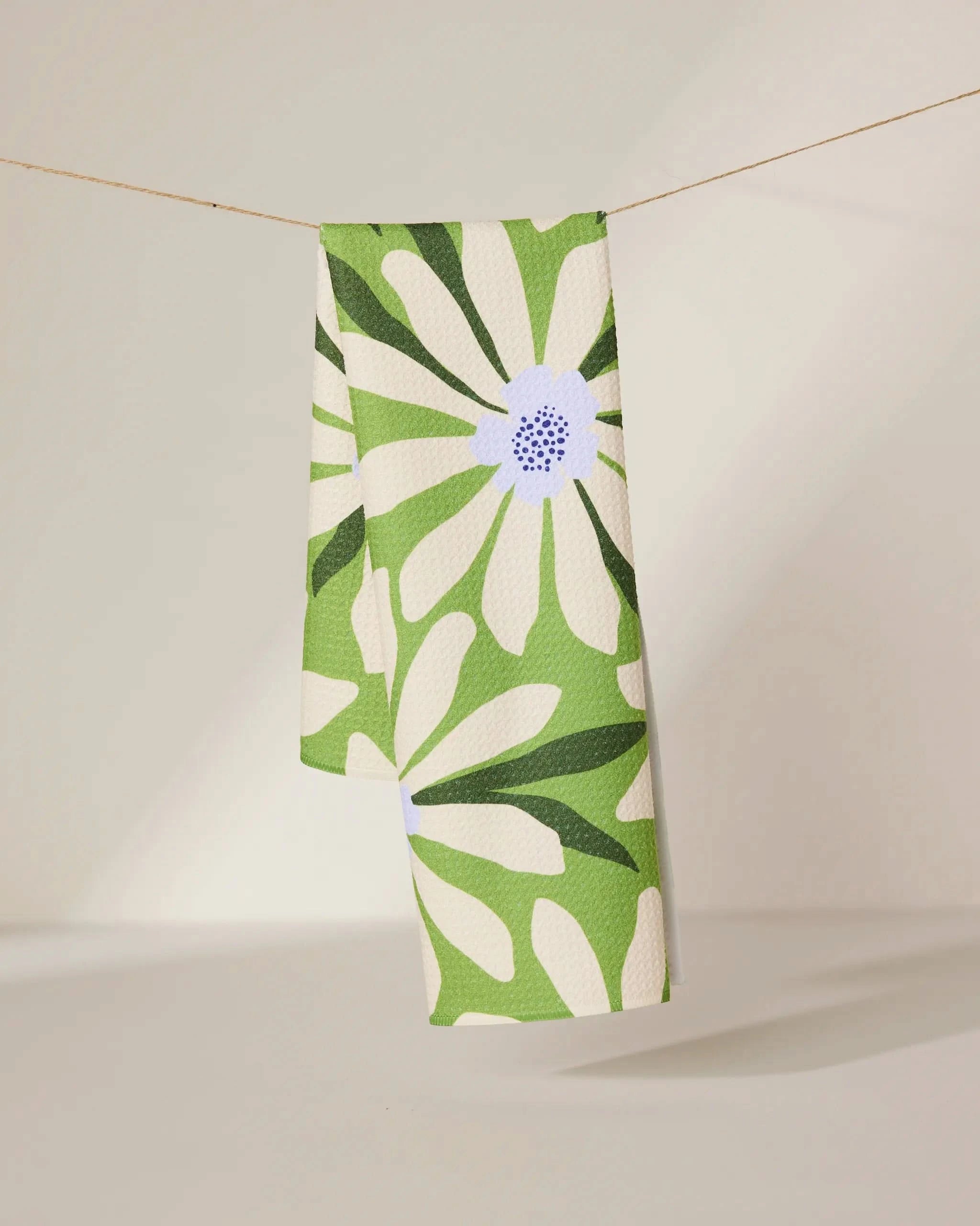 The GEOMETRY Bliss and Bloom tea towel, featuring a bold green and white daisy floral pattern, hangs neatly over a clothesline against a softly lit plain background.