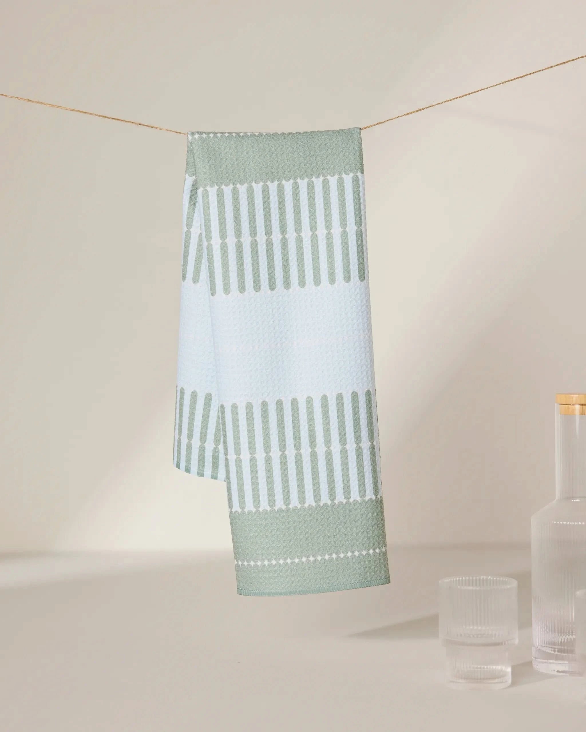 The Bâton Vert tea towel by GEOMETRY, featuring light blue and green patterns and made from recycled materials, hangs on a string with a clear glass bottle and two matching glasses nearby against a neutral background.