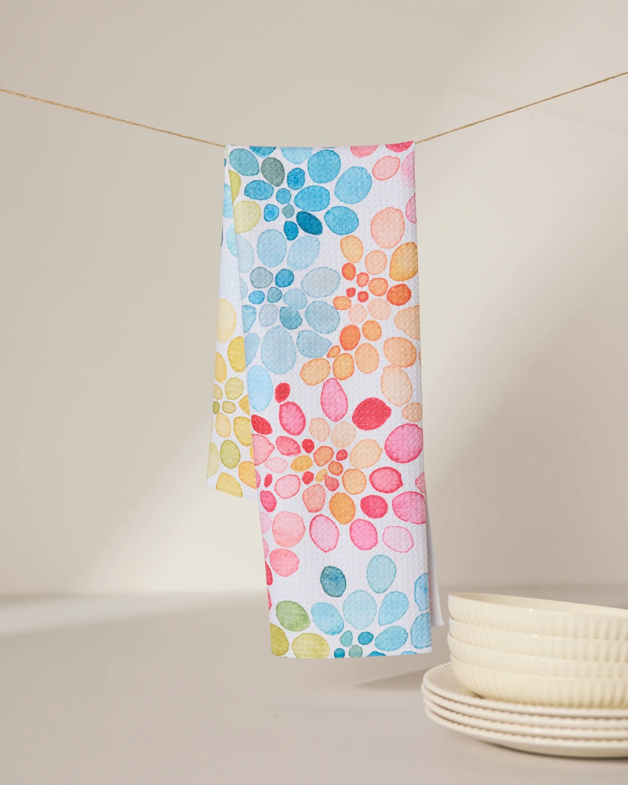 The GEOMETRY Bright Bouquet ultra-absorbent dish towel, featuring a vibrant floral petal design, hangs on a clothesline with cream-colored stacked bowls and plates in the foreground.