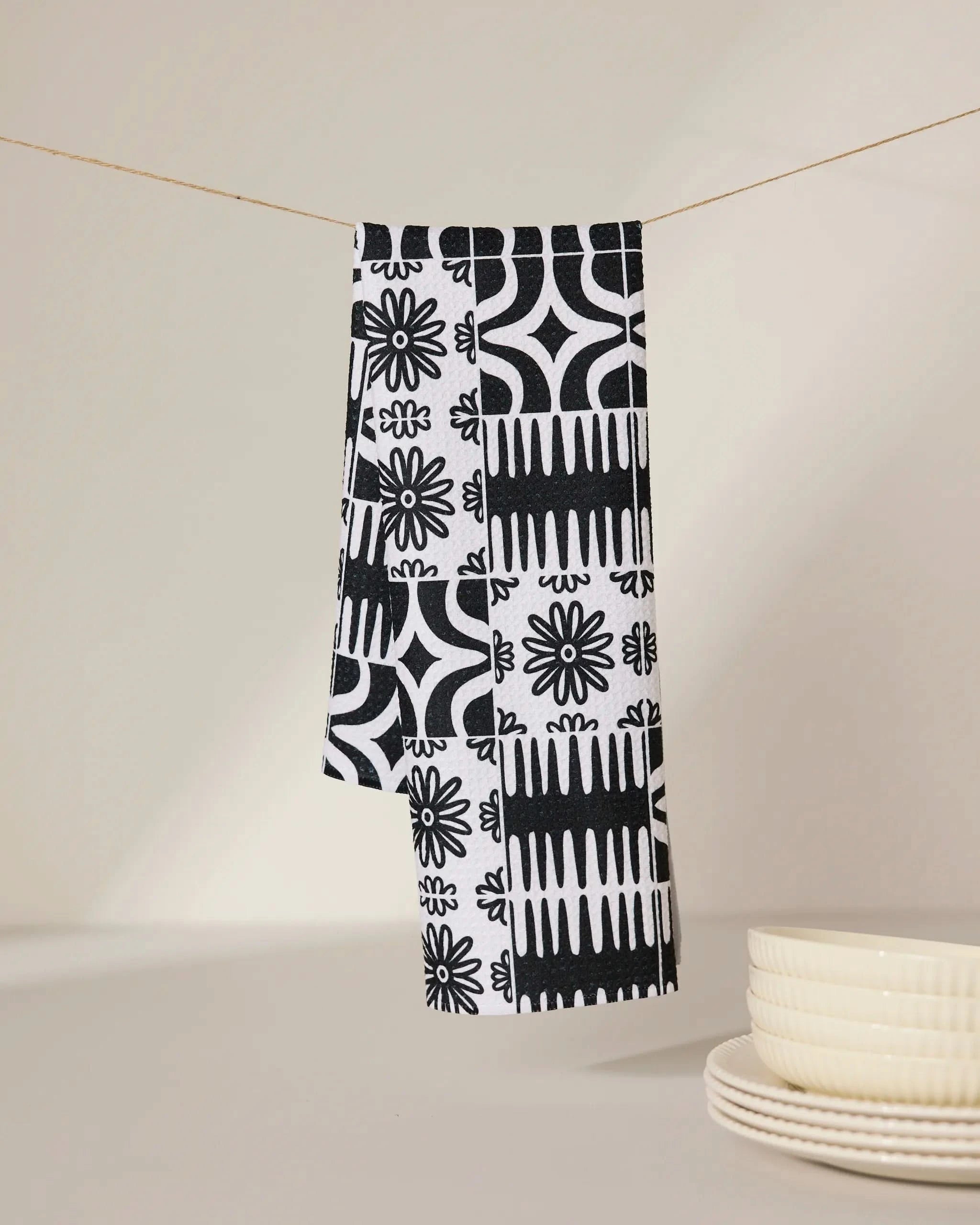 The GEOMETRY Bohemian Mosaic recycled kitchen towel, featuring a black and white pattern, hangs on a thin string above a beige surface, with a stack of white ceramic plates nearby.