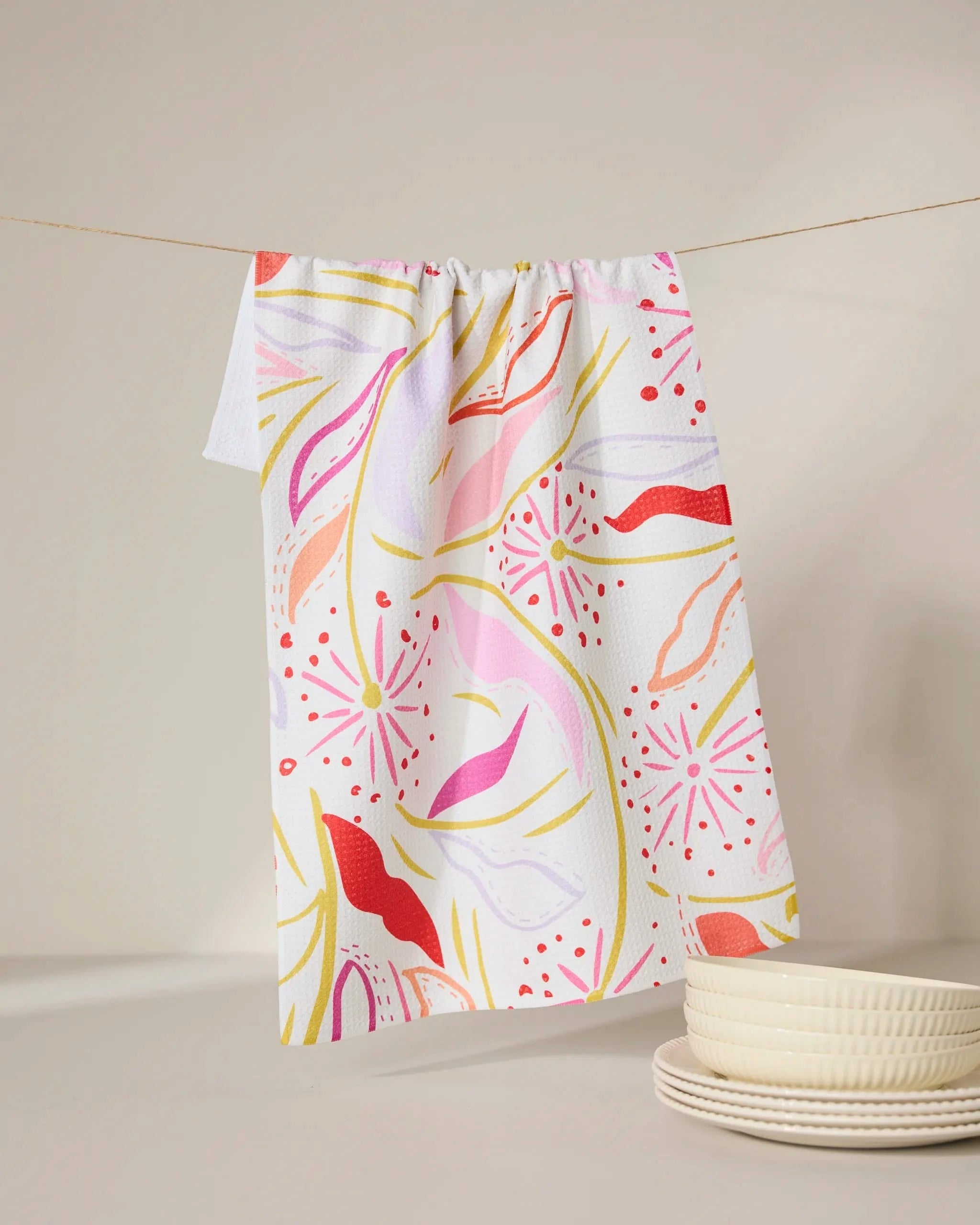 The GEOMETRY Grand Bloom kitchen towel, made from recycled materials, features vibrant pink, red, yellow, and orange patterns. Hanging on a line beside white textured plates on a softly lit surface, it brings color to any space.