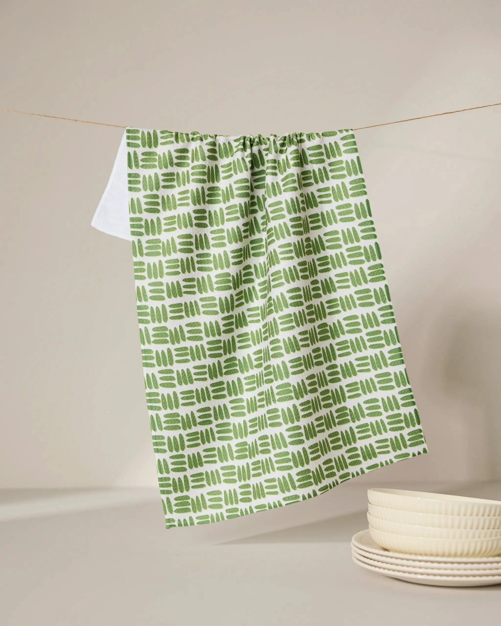The GEOMETRY Grass Hatch tea towel, ultra-absorbent and featuring a green and white pattern, hangs on a clothesline above a white surface next to a stack of cream-colored dishes.