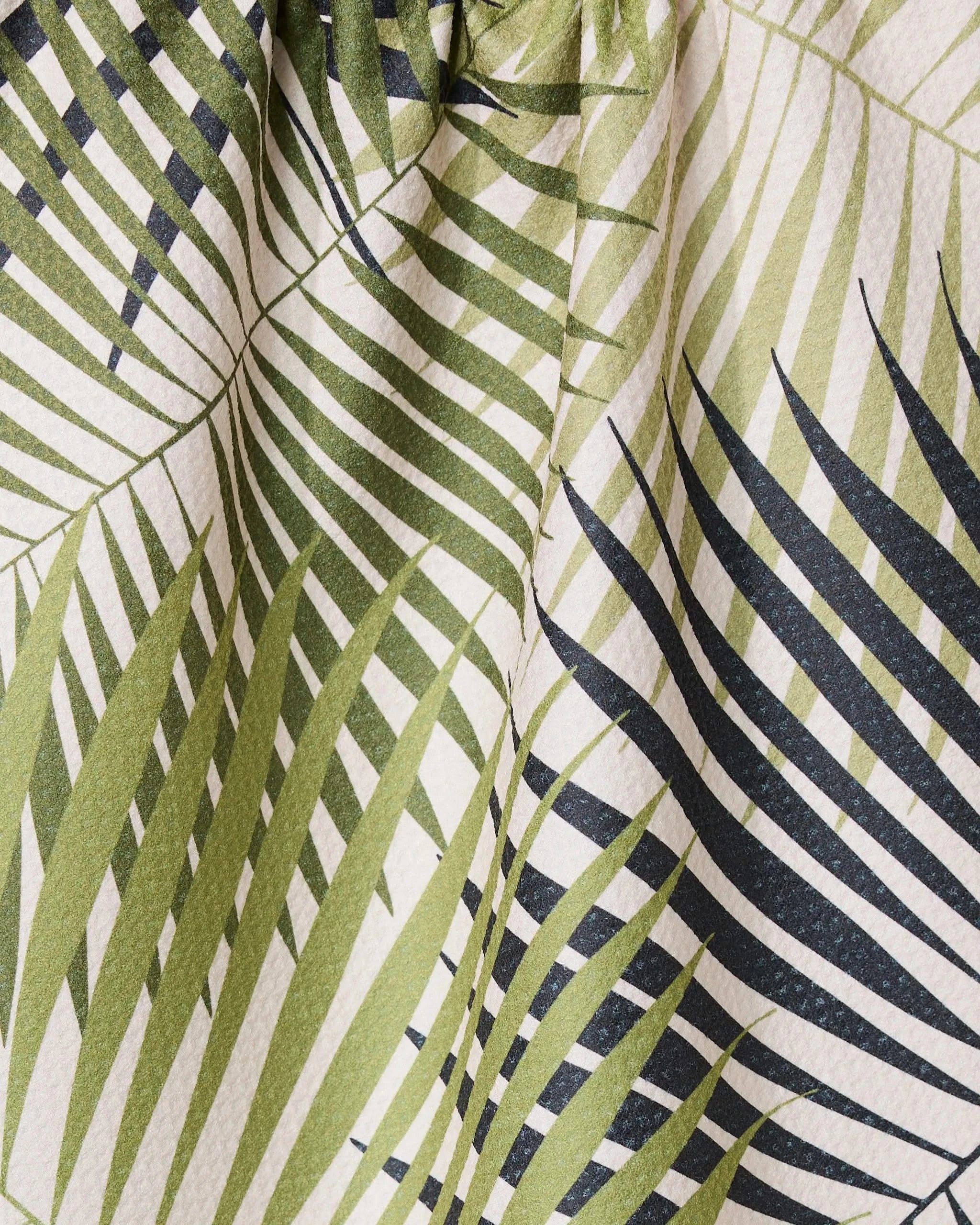 Close-up of GEOMETRY’s Green Fronds eco-friendly tea towel, featuring overlapping green palm leaves on a light background, with visible creases and textured material.