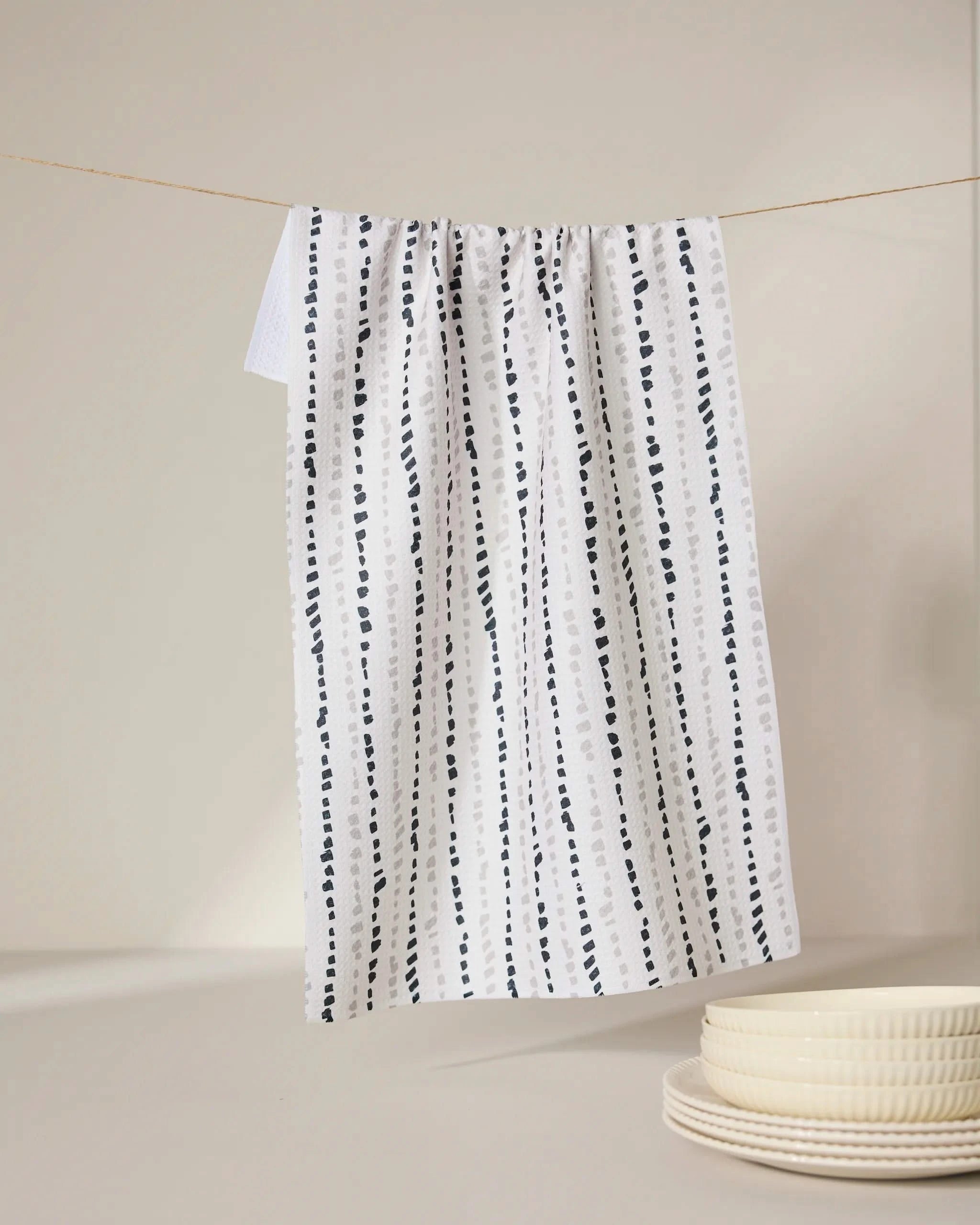A GEOMETRY Grey Prints recycled kitchen towel with vertical dotted lines hangs on a string, with stacked cream-colored plates in the foreground on a neutral background.