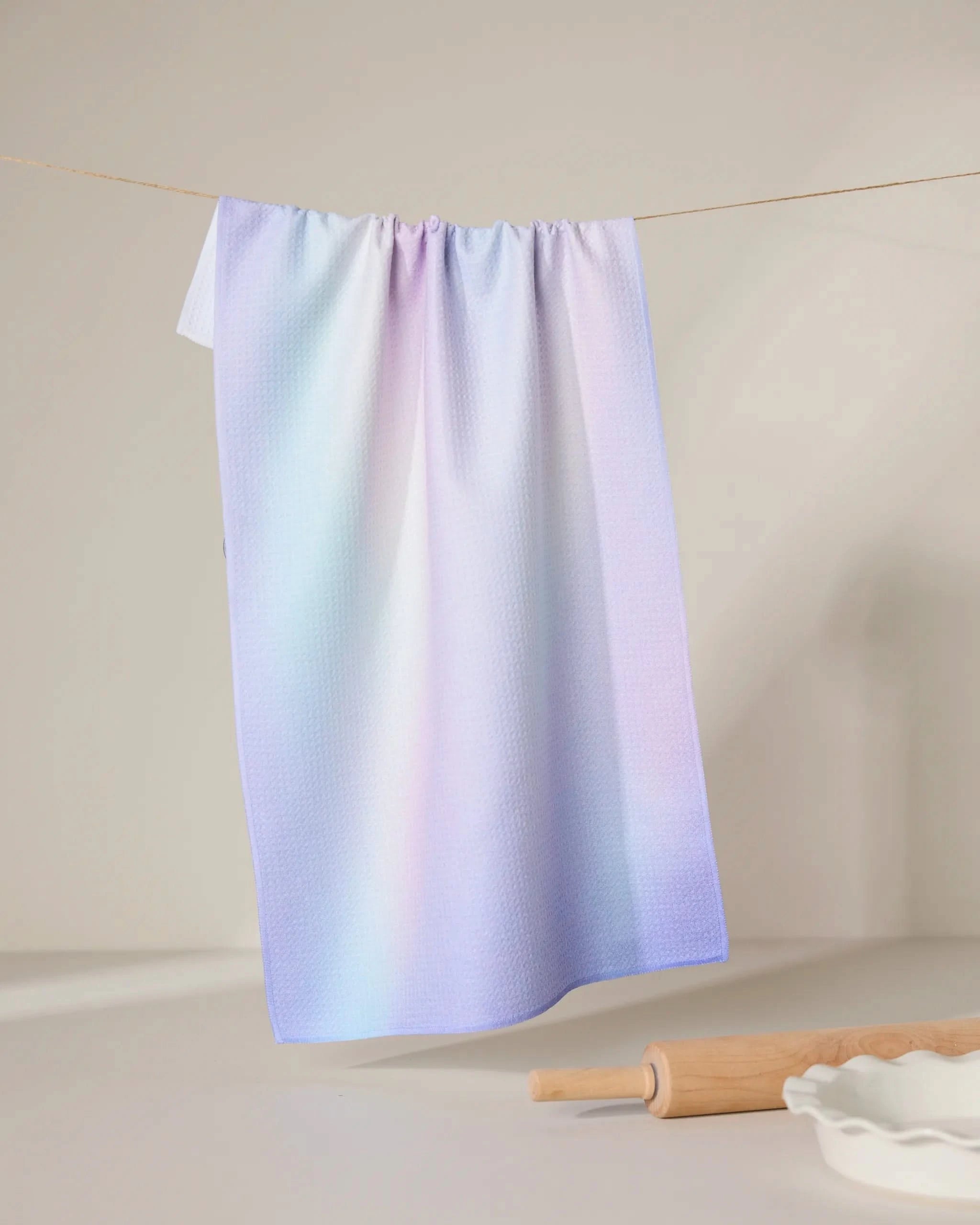 The GEOMETRY Iridescence kitchen towel, featuring an ultra-absorbent pastel gradient in pink, blue, and purple, hangs on a line. Below are a wooden rolling pin and a white fluted dish on a light surface.