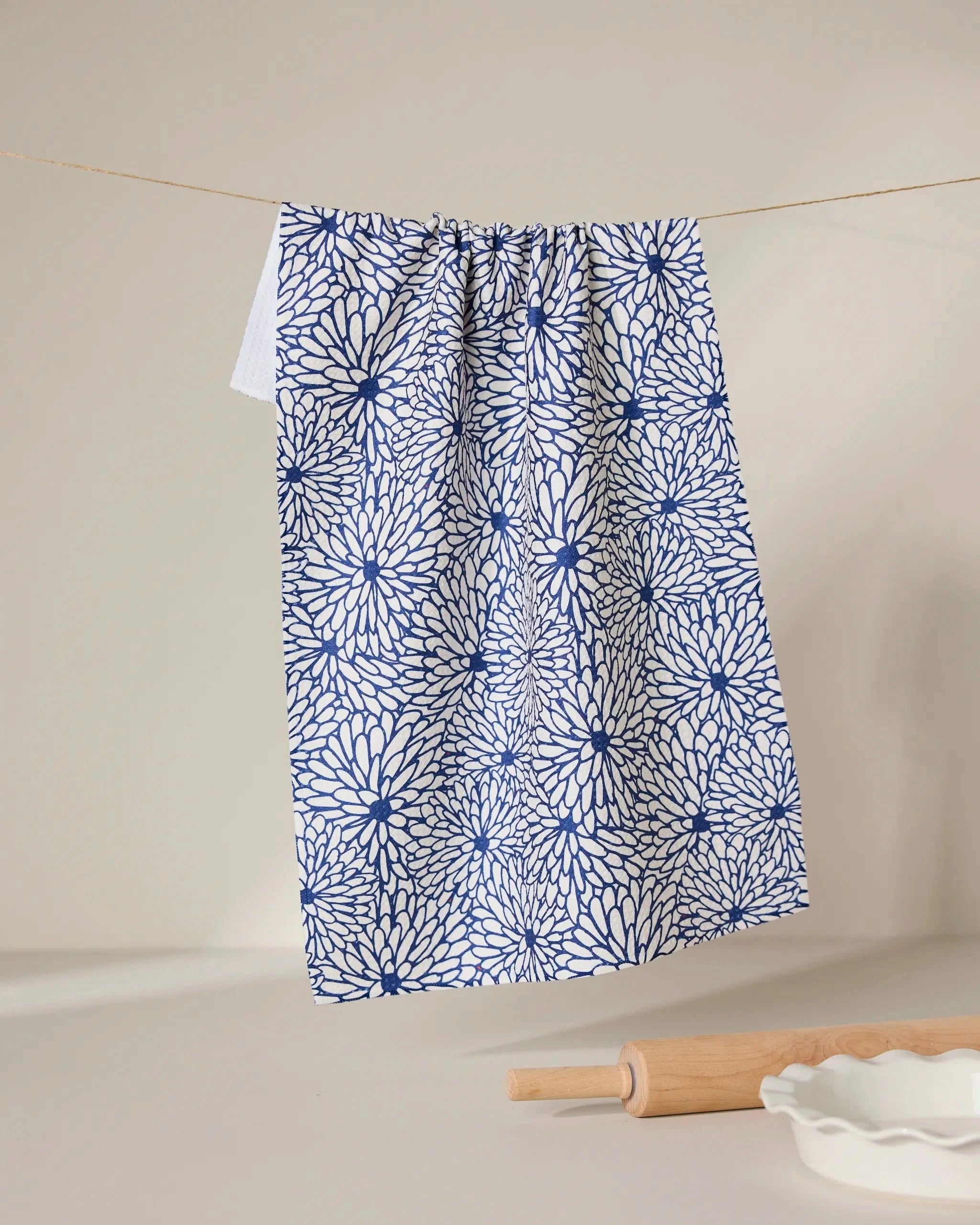 The GEOMETRY Jar Jar tea towel, featuring a blue and white floral design and crafted with Geoweave technology for superior absorbency, hangs on a clothesline. Below, a wooden rolling pin and a white fluted dish sit on a light surface.