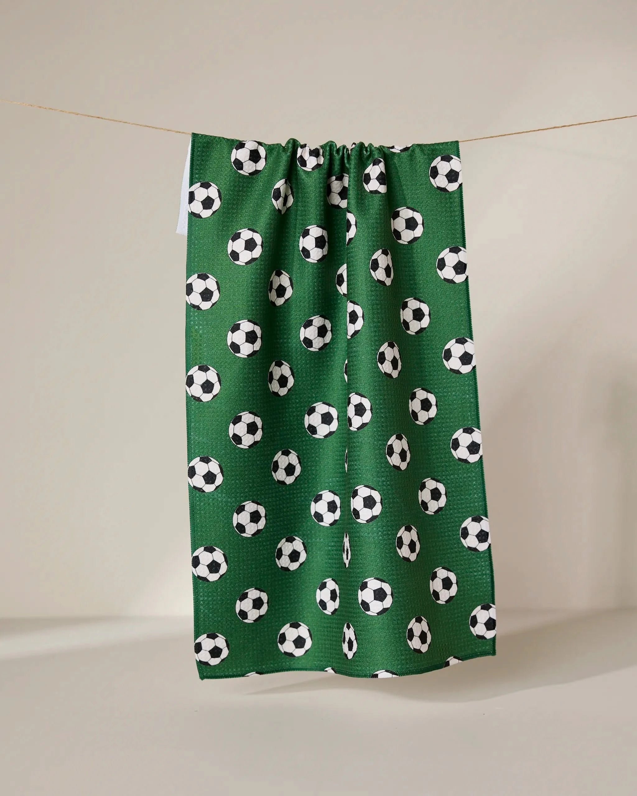 The GEOMETRY Kick Off eco-friendly kitchen towel in green features a repeating black and white soccer ball pattern and hangs on a line against a plain, light-colored background.
