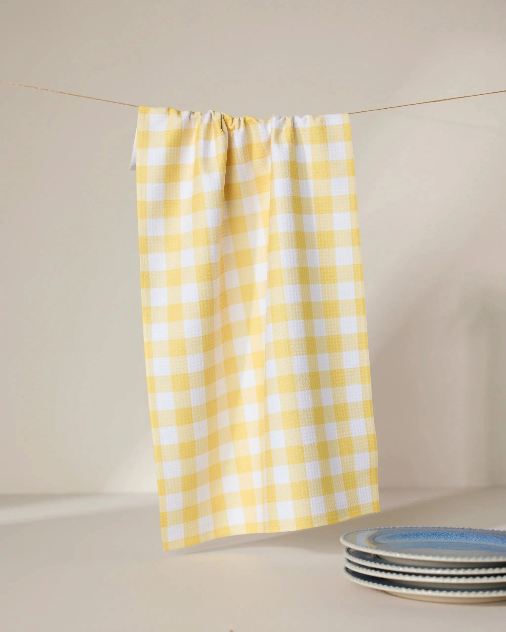 The GEOMETRY Lemon Gingham recycled kitchen towel hangs on a line with three blue and white patterned ceramic plates stacked nearby, all set against a neutral background.