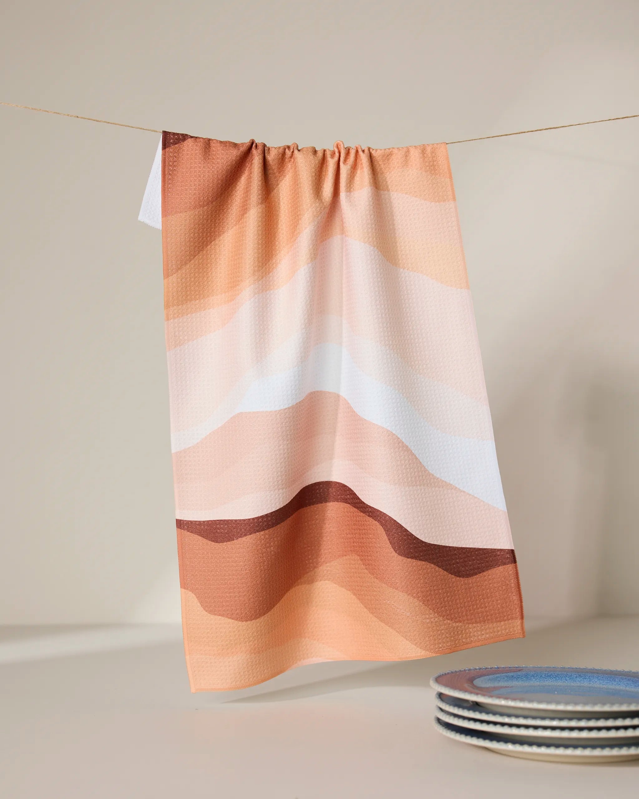 The GEOMETRY Levels of Fall kitchen tea towel, featuring an abstract wavy pattern in peach, tan, and brown and made with super-absorbent Geoweave technology, hangs above a white surface near stacked ceramic plates with colorful rims.
