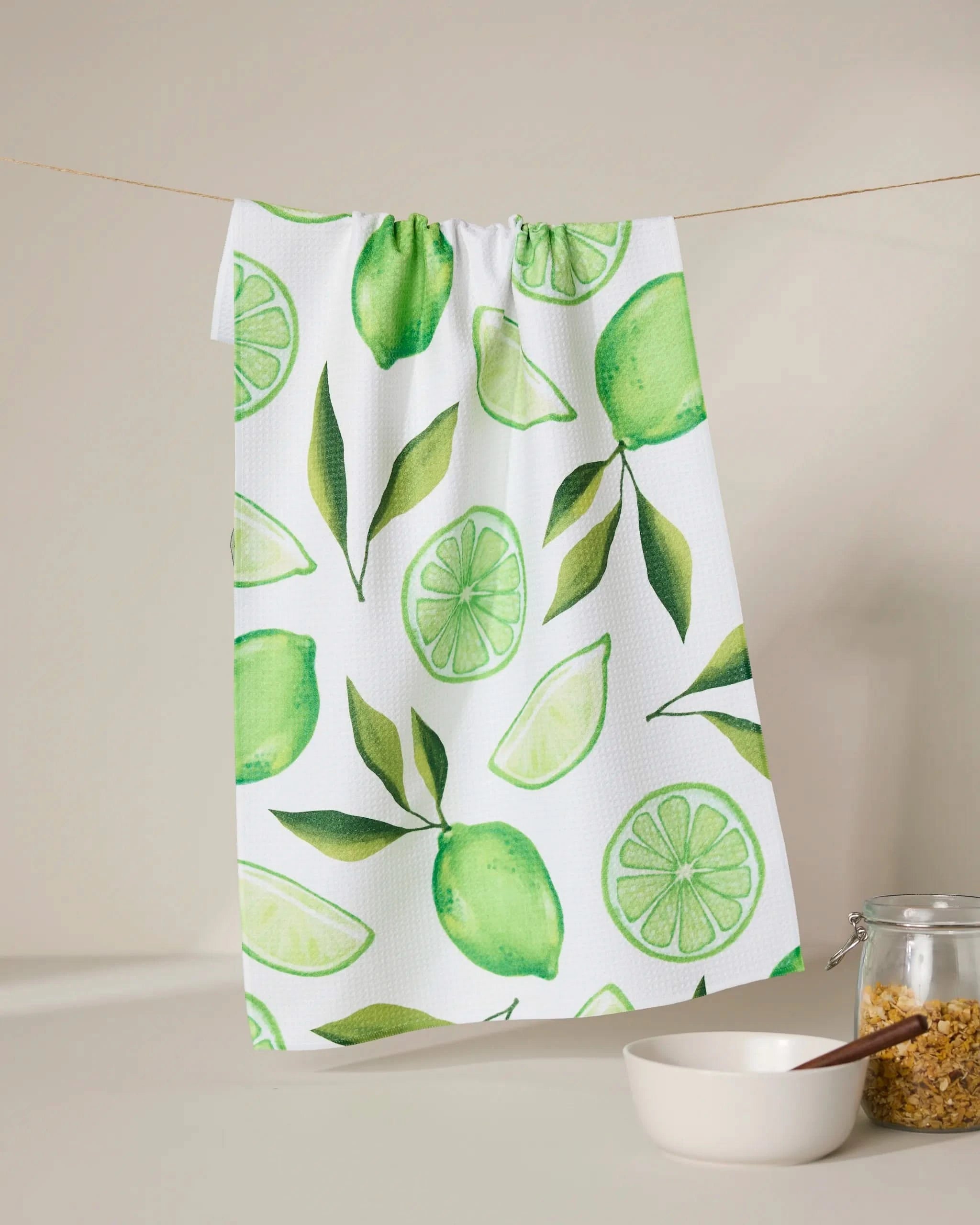 The GEOMETRY Lima kitchen towel, made from recycled materials and featuring green lime and leaf designs, hangs above a glass jar of brown sugar and a white bowl with a wooden spoon.