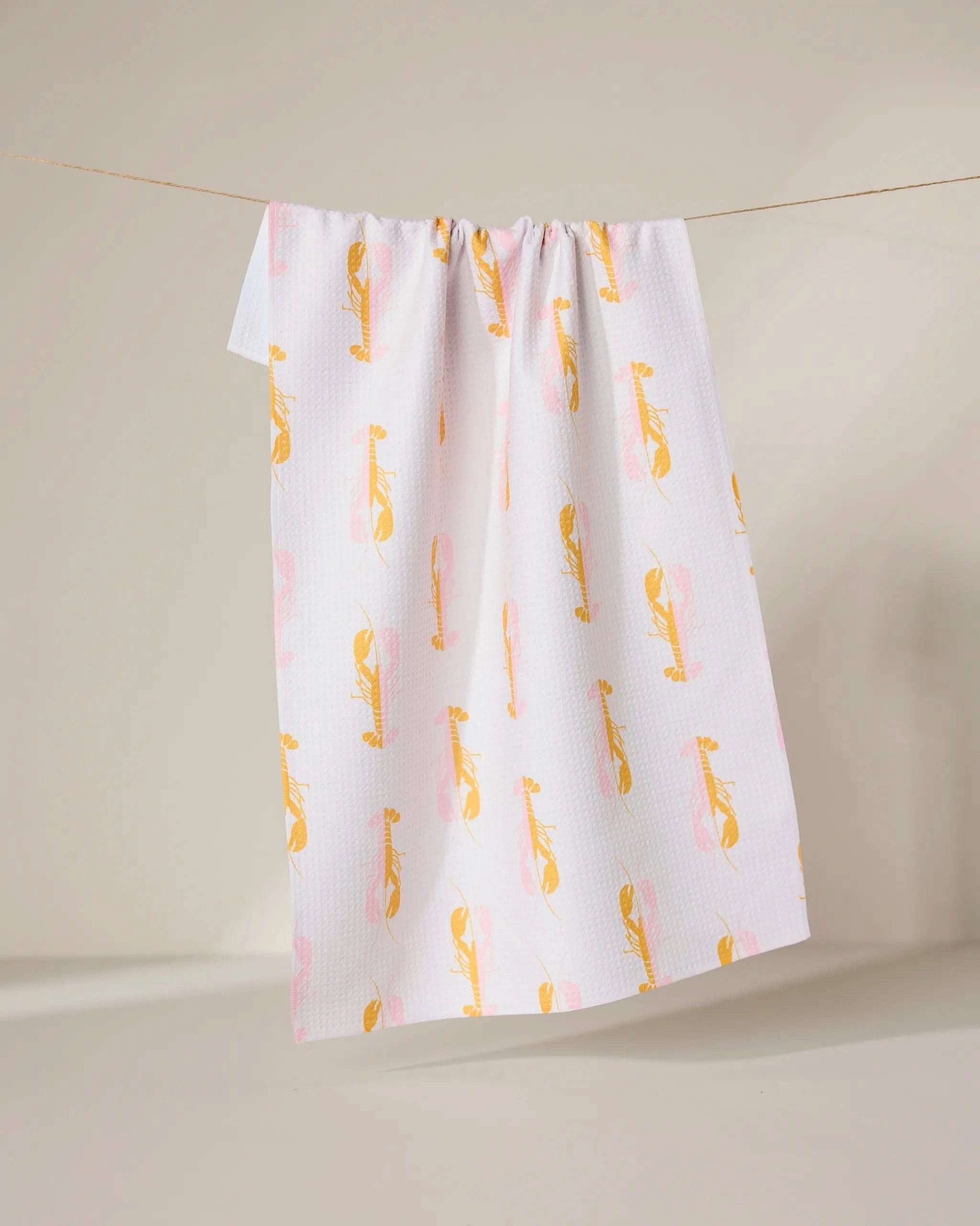 The GEOMETRY Lobster Love towel, made from recycled materials, features a white background with a yellow and pink abstract bird pattern and hangs on a clothesline against a neutral backdrop.