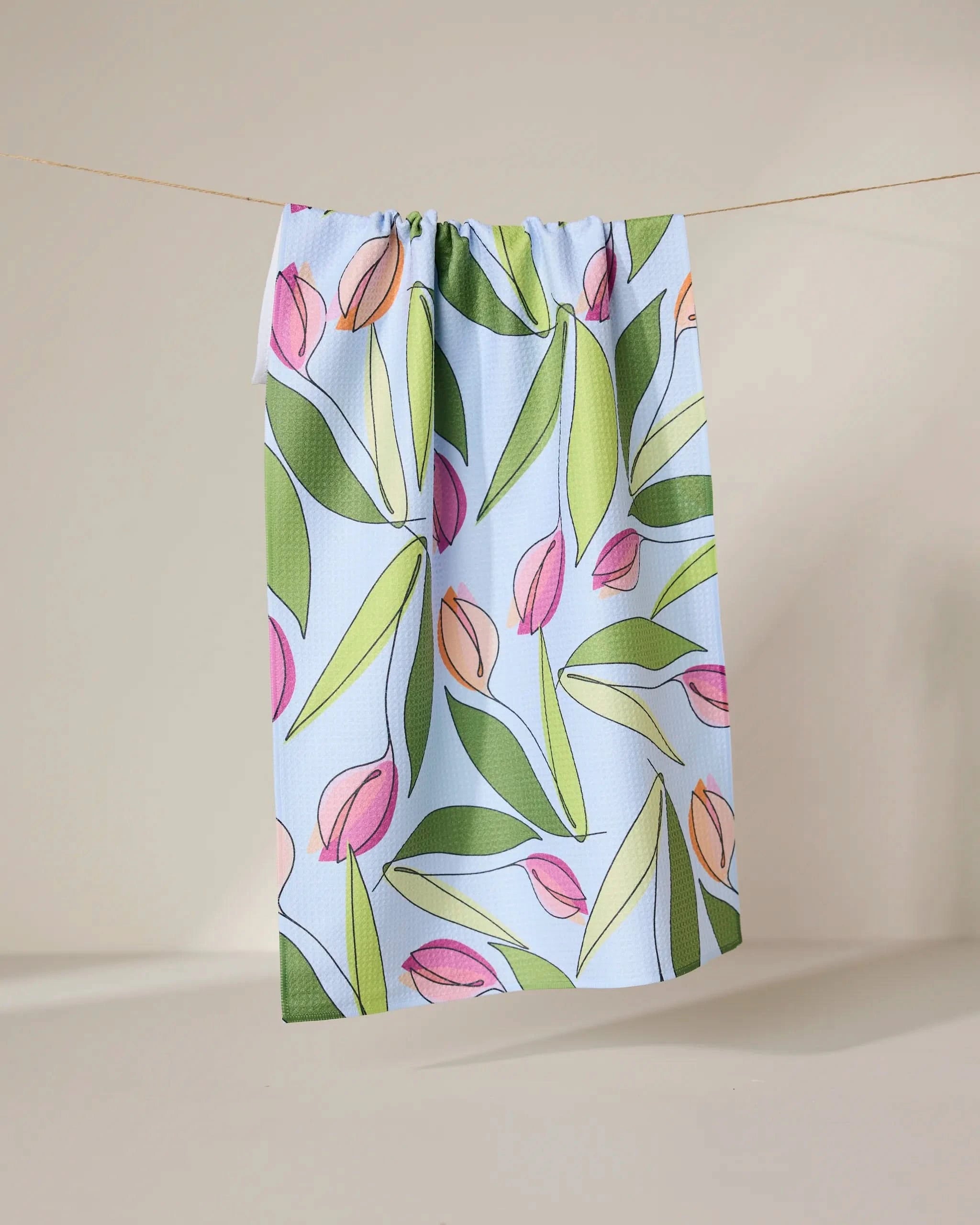 The GEOMETRY May Flowers recycled kitchen towel, featuring a pink and green tulip and leaf pattern on light blue, hangs from a string against a neutral backdrop.