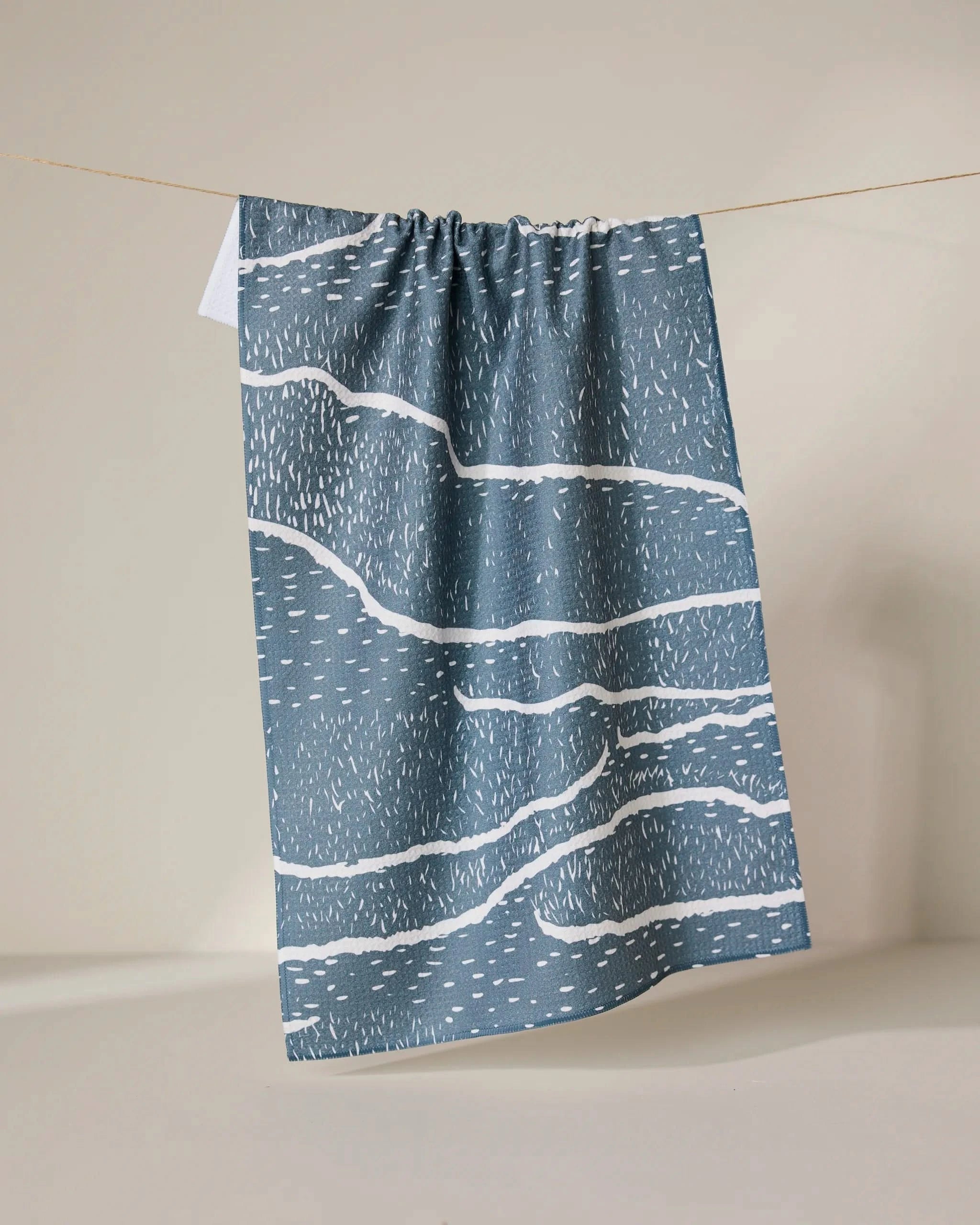 The GEOMETRY Wetlands - Midnight tea towel, featuring abstract wavy white lines and dashes on a blue base, hangs from a string against a neutral background. This absorbent dish towel combines style and functionality.