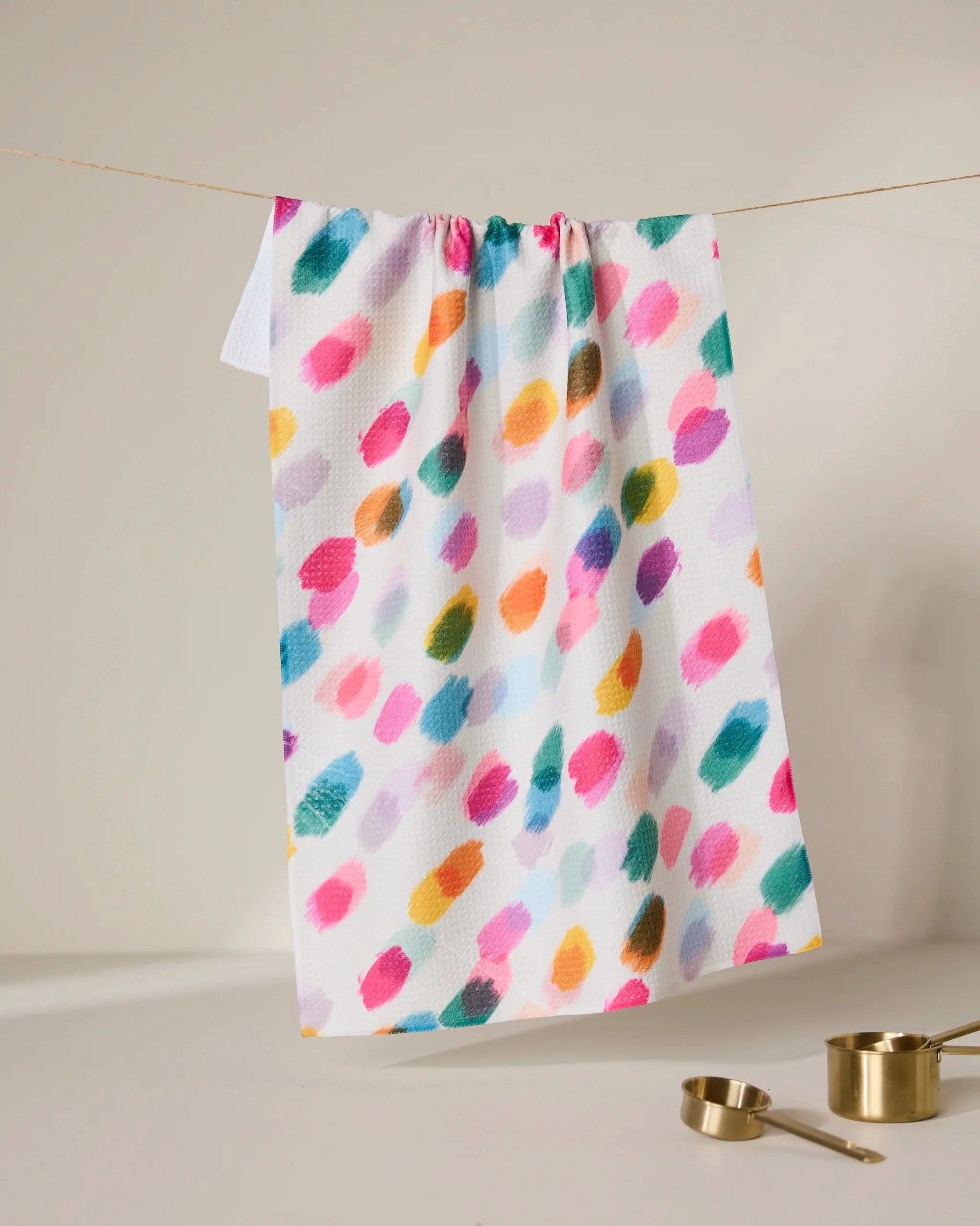 The GEOMETRY Downpour tea towel, made from recycled materials and featuring colorful, irregular brushstrokes, hangs on a softly lit line. Below, gold measuring cups and spoons rest on a neutral surface. Ultra-absorbent design.