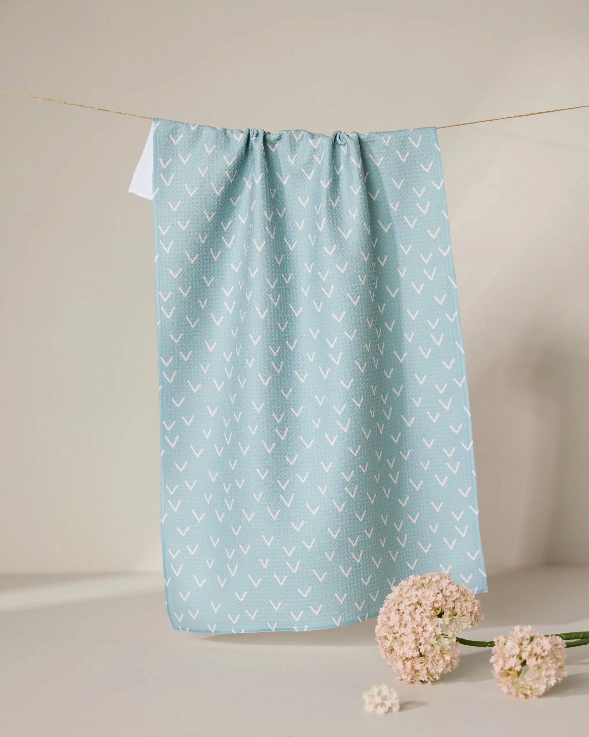 The GEOMETRY Drawn Doves tea towel, featuring a quick-drying light blue fabric with a white arrow pattern, hangs on a clothesline. Below it, light pink flowers are arranged on a beige surface against a neutral backdrop.