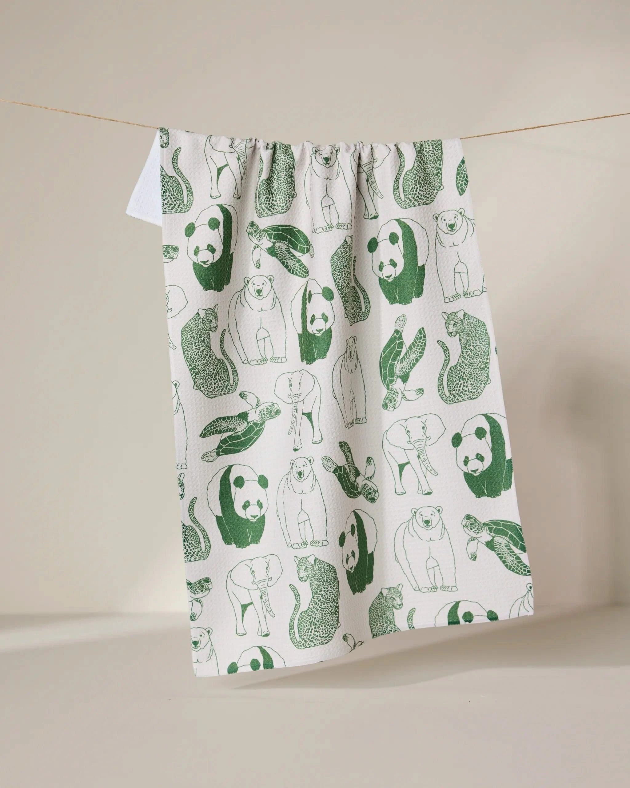 The GEOMETRY Endangered tea towel, made from recycled materials, dries quickly and features green illustrations of pandas, snakes, rabbits, and otters. It hangs on a clothesline against a neutral background.