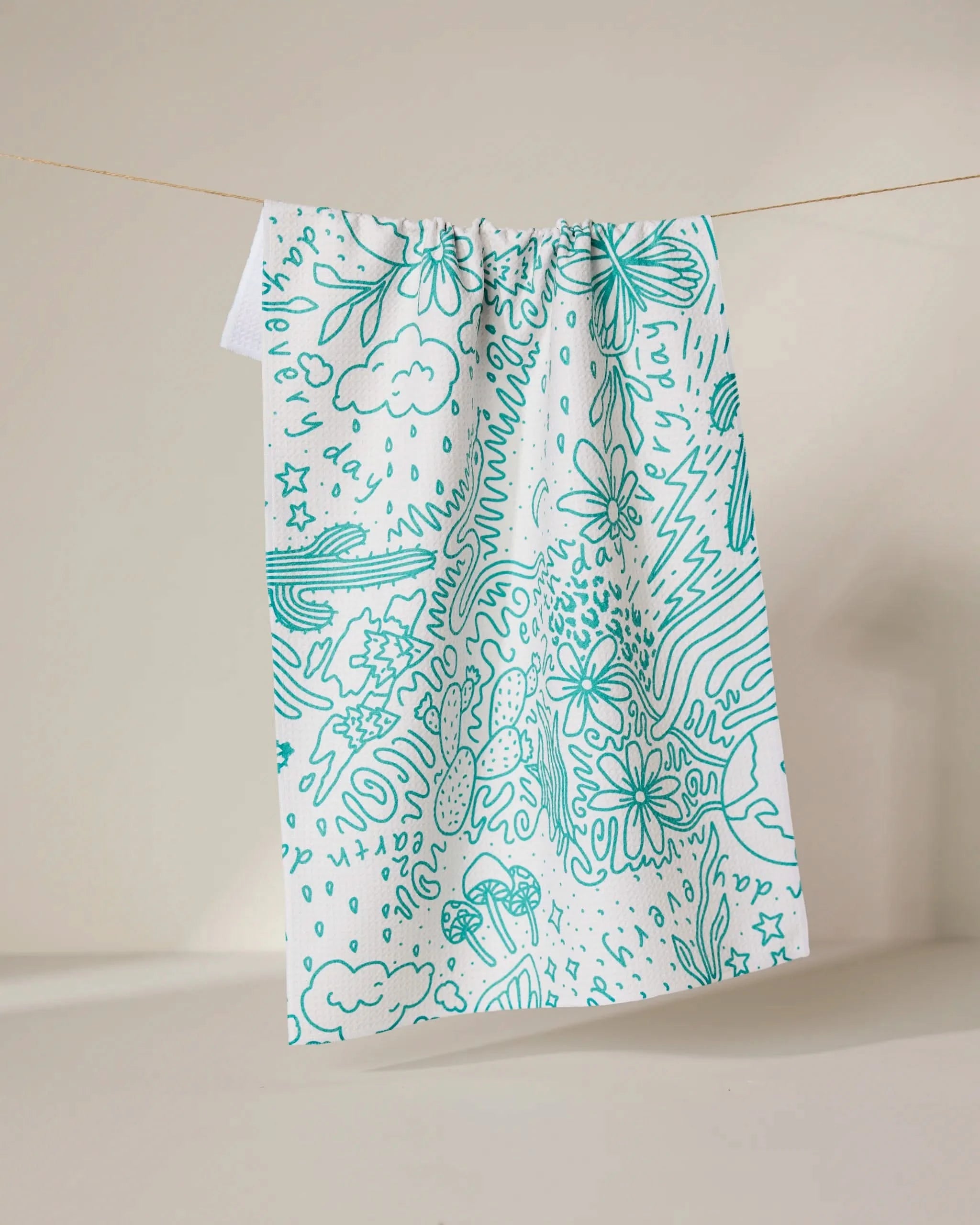 The Every Day tea towel by GEOMETRY, made from recycled materials, displays teal line art of flowers, mushrooms, clouds, stars, and abstract shapes on white fabric, shown hanging against a neutral backdrop.