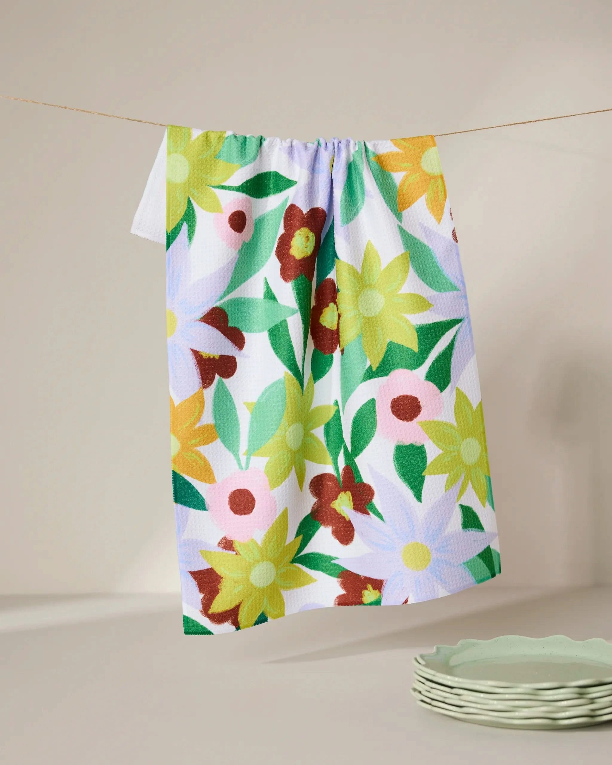 The GEOMETRY Floral Fields tea towel, featuring a vibrant yellow, green, red, and white floral design, hangs on a clothesline above pale green plates set against a neutral background.