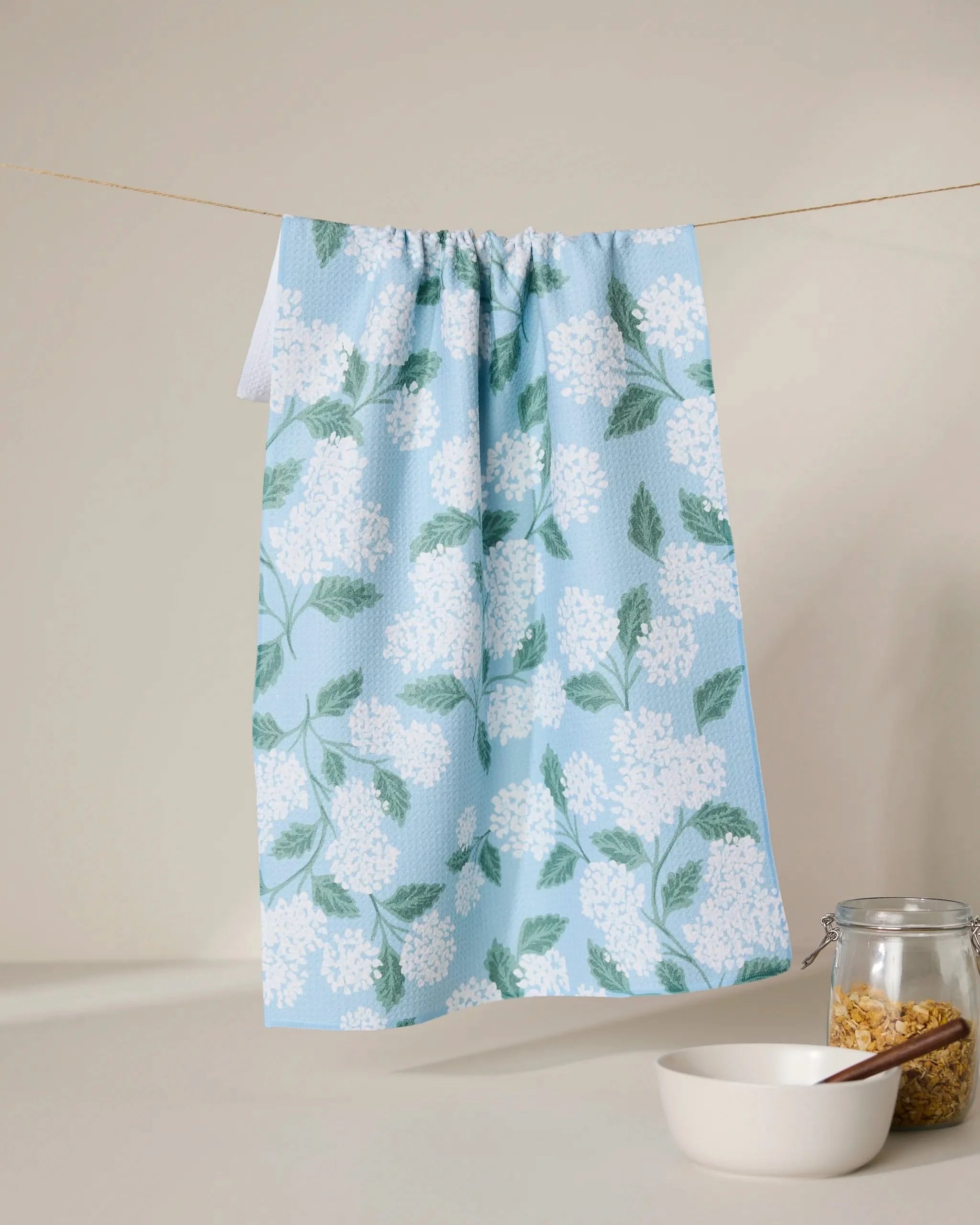 The GEOMETRY Garden Of Edith blue tea towel, featuring a white floral and green leaf pattern, hangs on a string above a white bowl and pasta-filled jar. Made from quick-drying, recycled materials.
