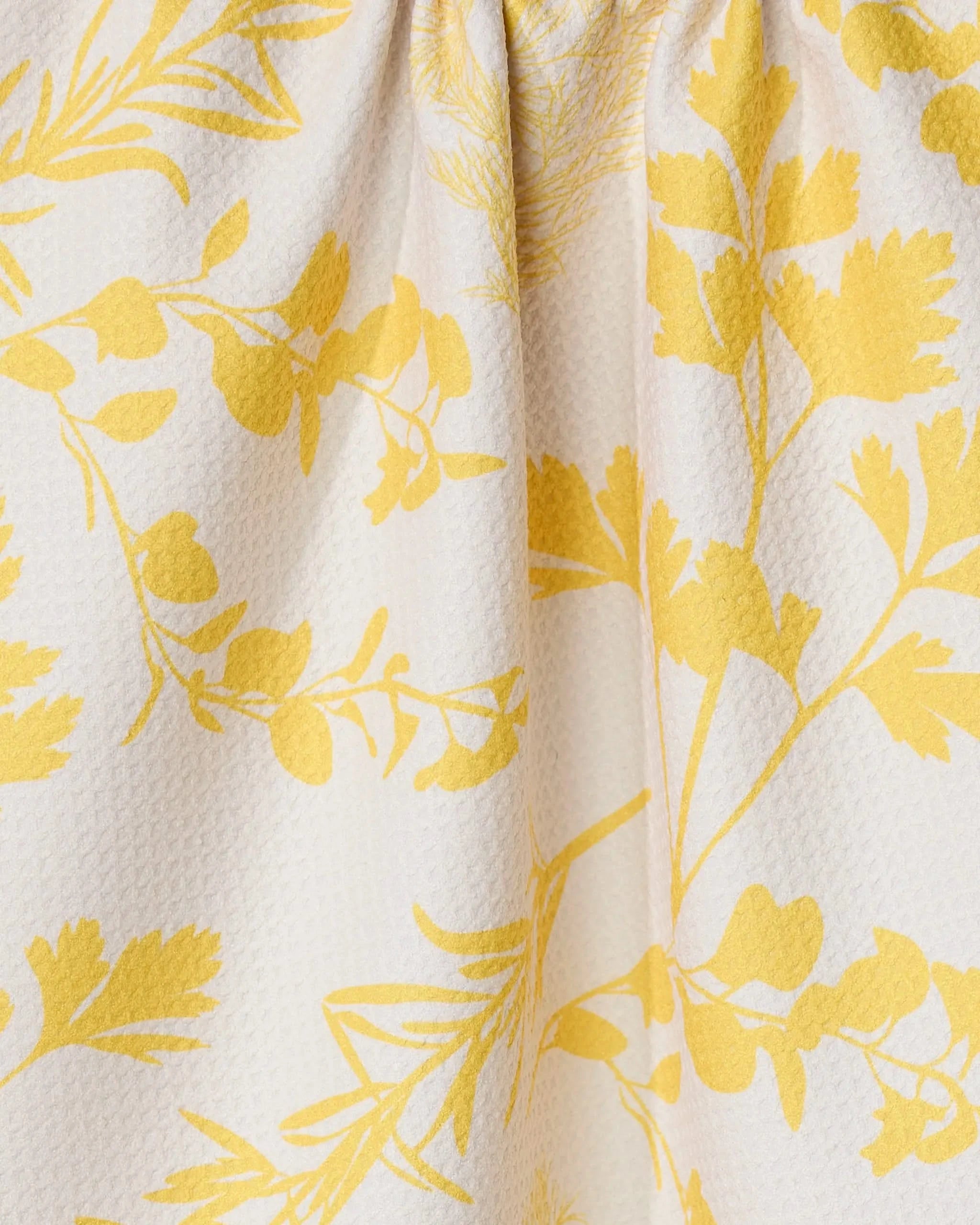 The GEOMETRY Garden of Gold tea towel showcases a close-up of its textured white fabric, adorned with a yellow botanical pattern of leaves and branches. Ultra-absorbent material is softly gathered at the top, creating gentle folds.