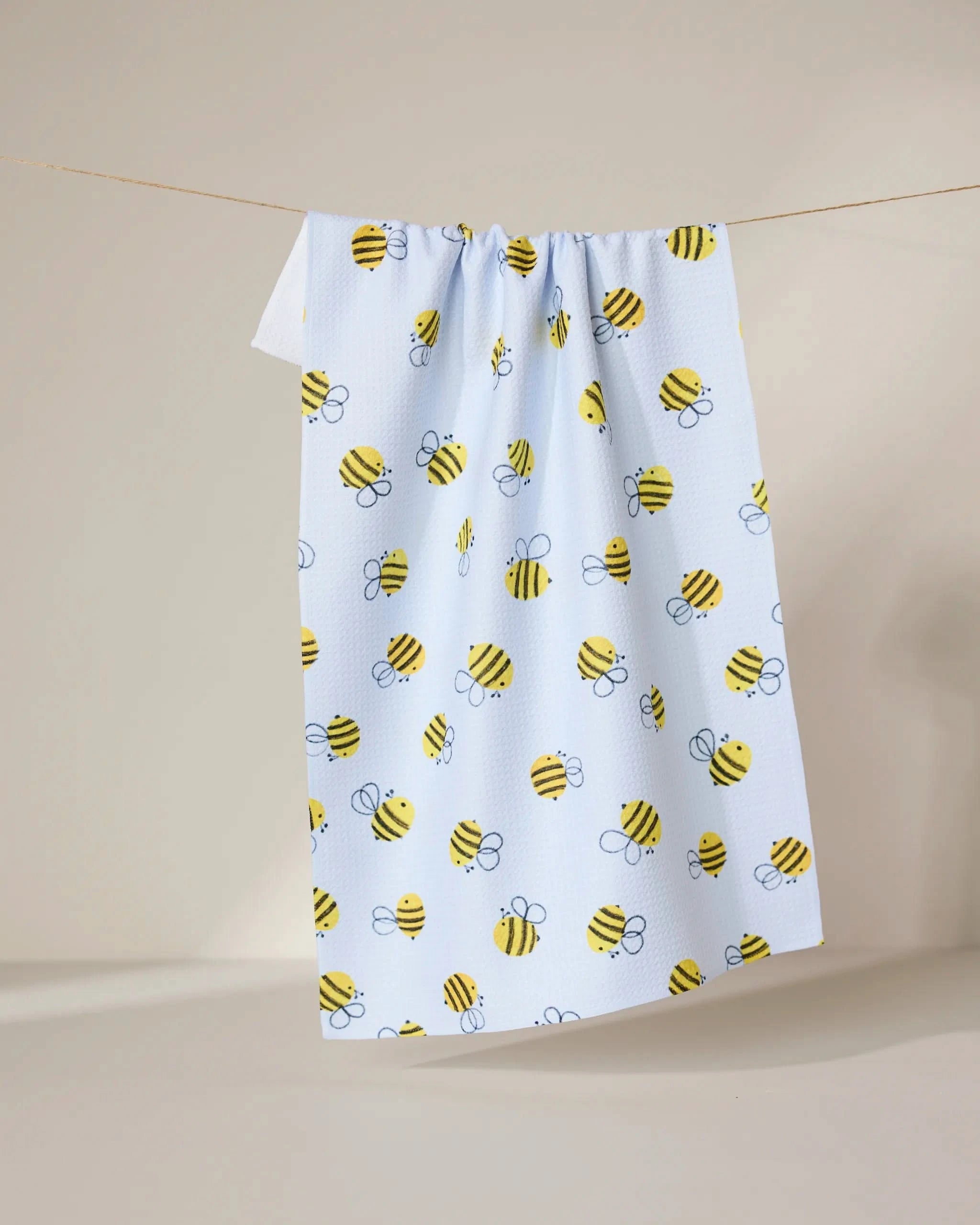 The GEOMETRY Buzzing tea towel, in light blue with a playful yellow and black bumblebee pattern, hangs on a string against a plain, light background.