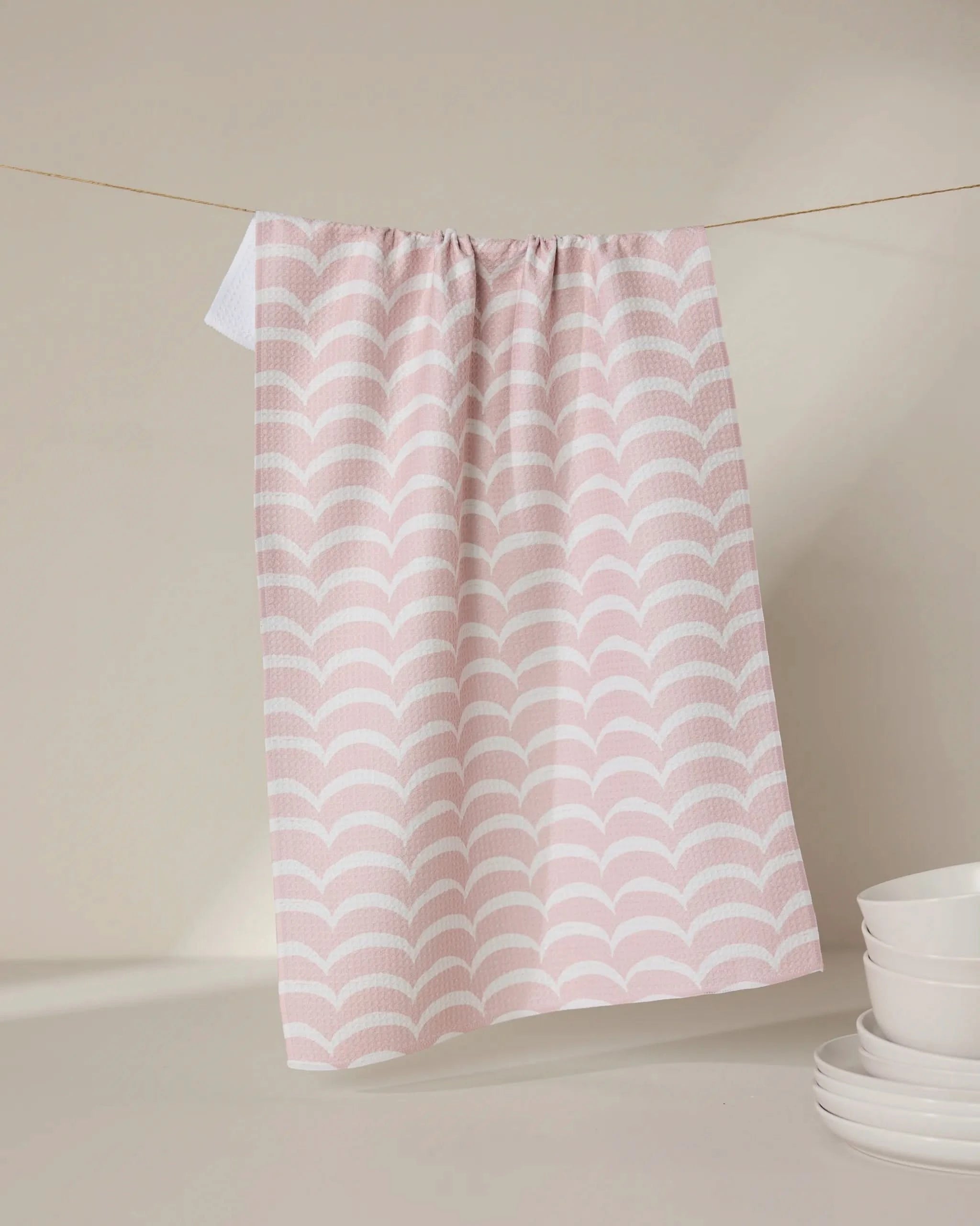 The GEOMETRY Cascading Pink eco-friendly kitchen towel with a scallop pattern hangs on a clothesline, while white ceramic bowls and saucers are stacked nearby on a beige surface against a neutral background.