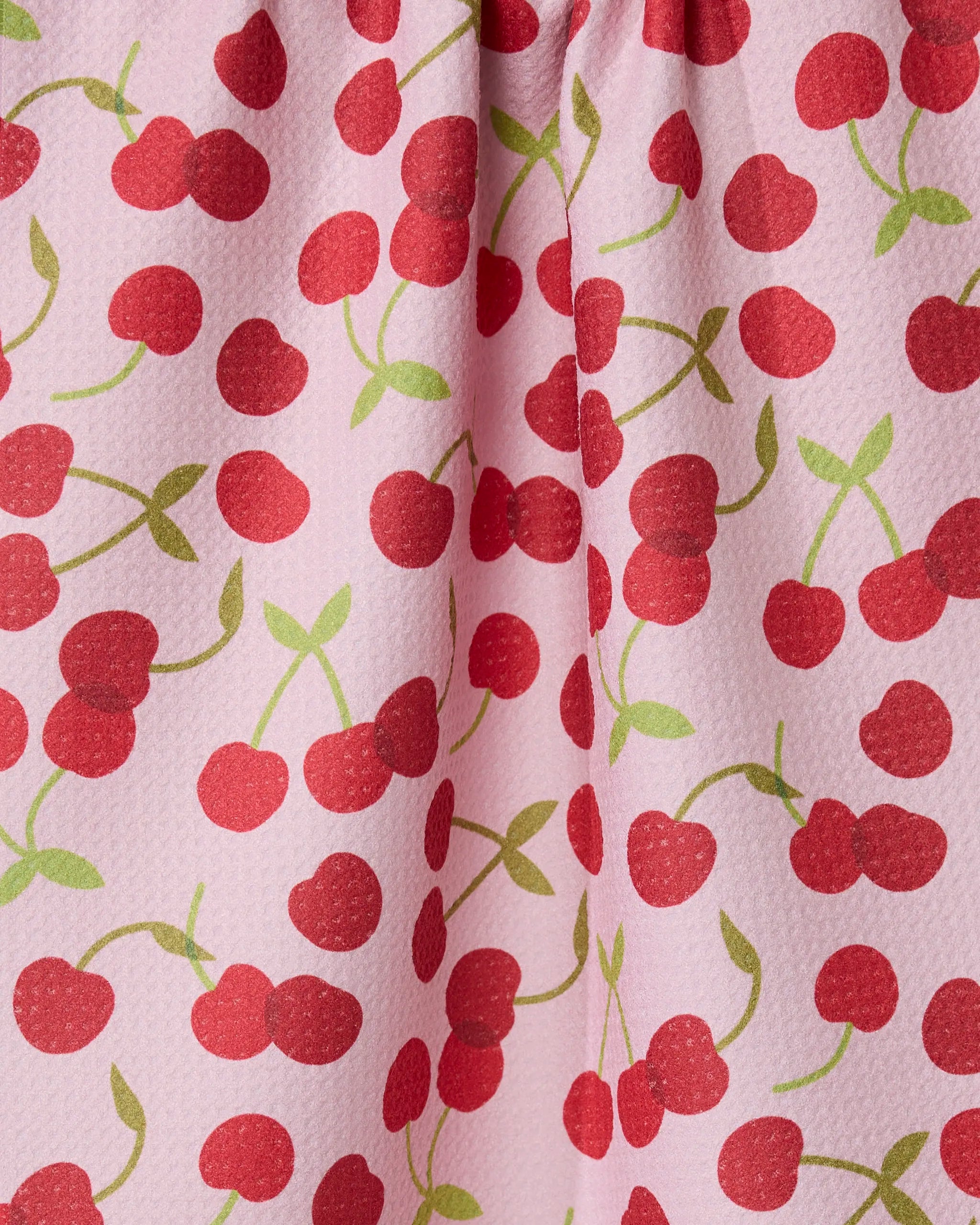 A close-up of Cheery Cherries by GEOMETRY shows pink fabric with red cherries and green stems, a lightly textured surface, and gentle folds—ideal for a recycled kitchen towel or ultra-absorbent tea towel.