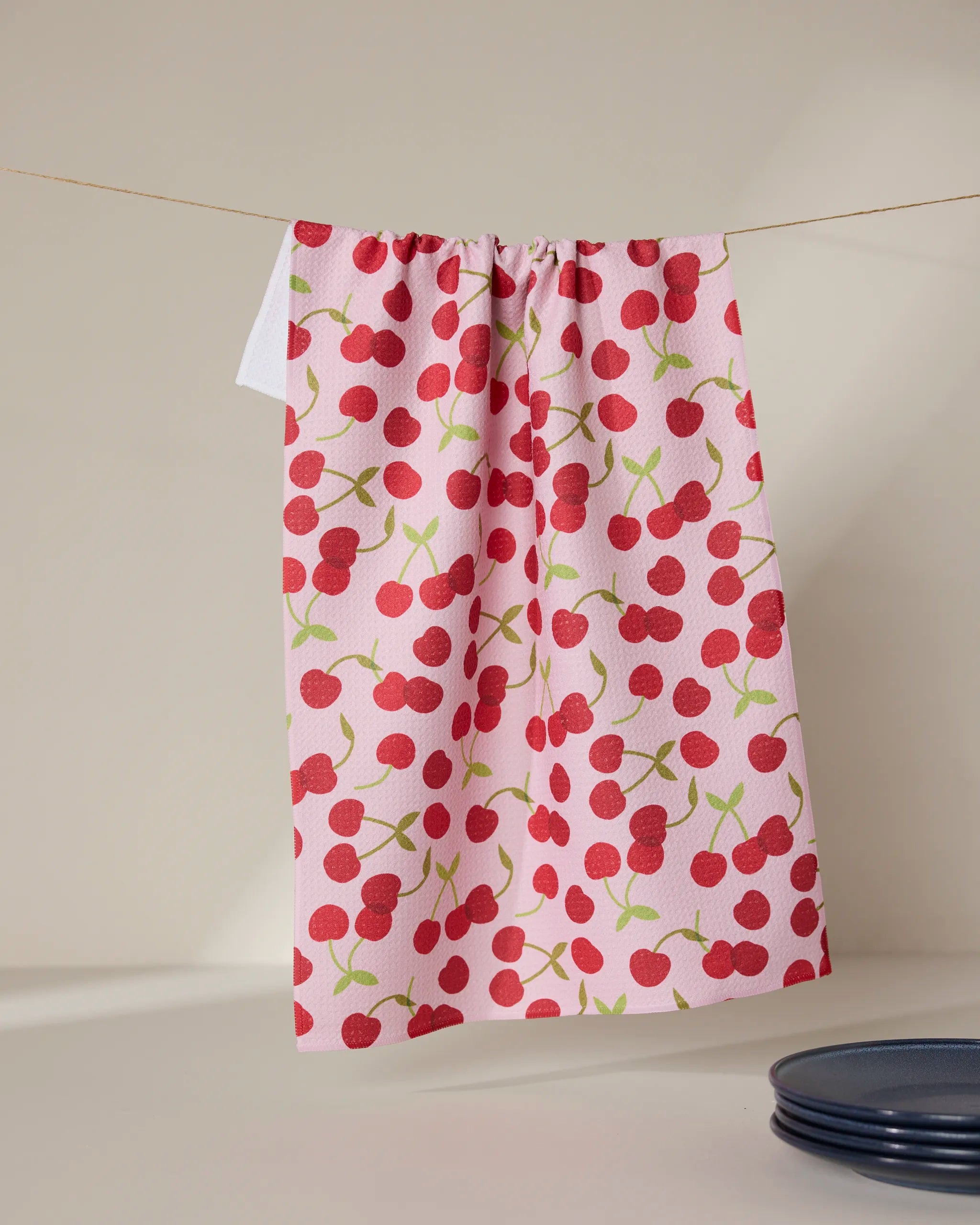 The Cheery Cherries towel by GEOMETRY, featuring a pink ultra-absorbent fabric with a bright red cherry pattern, hangs on a line against a neutral background, while a dark blue plate appears at the bottom right.