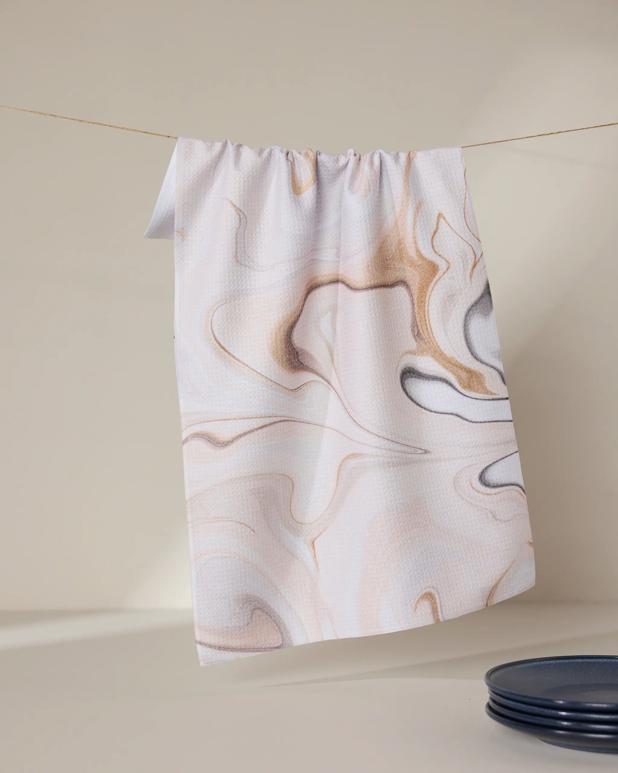 The GEOMETRY Choco tea towel, featuring a light brown and gray marbled pattern, hangs on a clothesline against a beige backdrop with a dark blue plate partly visible at the bottom right.