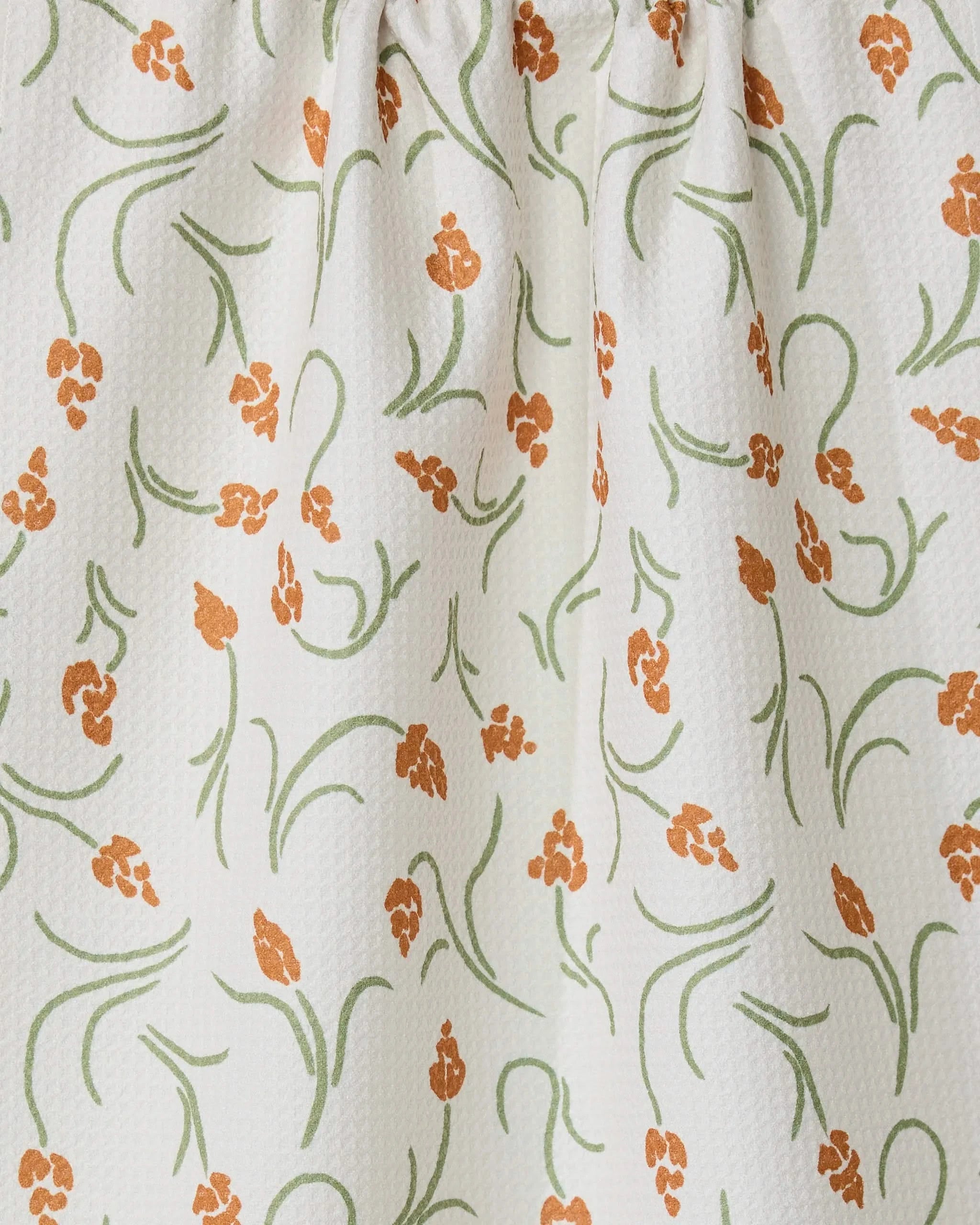 The GEOMETRY Crayon Floral tea towel features white textured fabric with orange floral clusters and green curving stems and leaves. Slightly gathered at the top, it offers style, function, and a light, airy look.