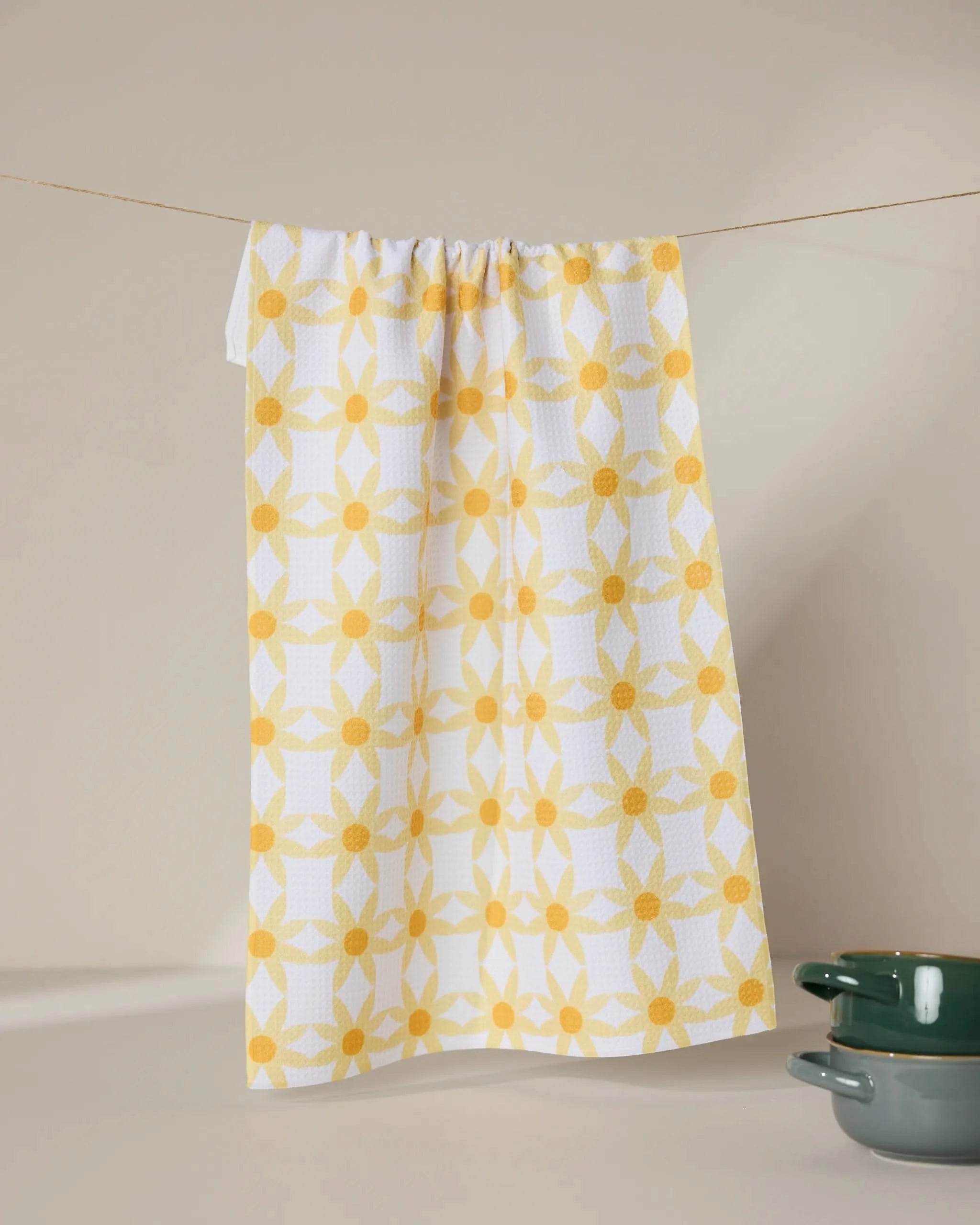 The GEOMETRY Daisies tea towel, featuring a yellow geometric floral pattern and made from recycled materials, hangs on a string. Two green ceramic bowls are stacked in the bottom right corner against a neutral background.