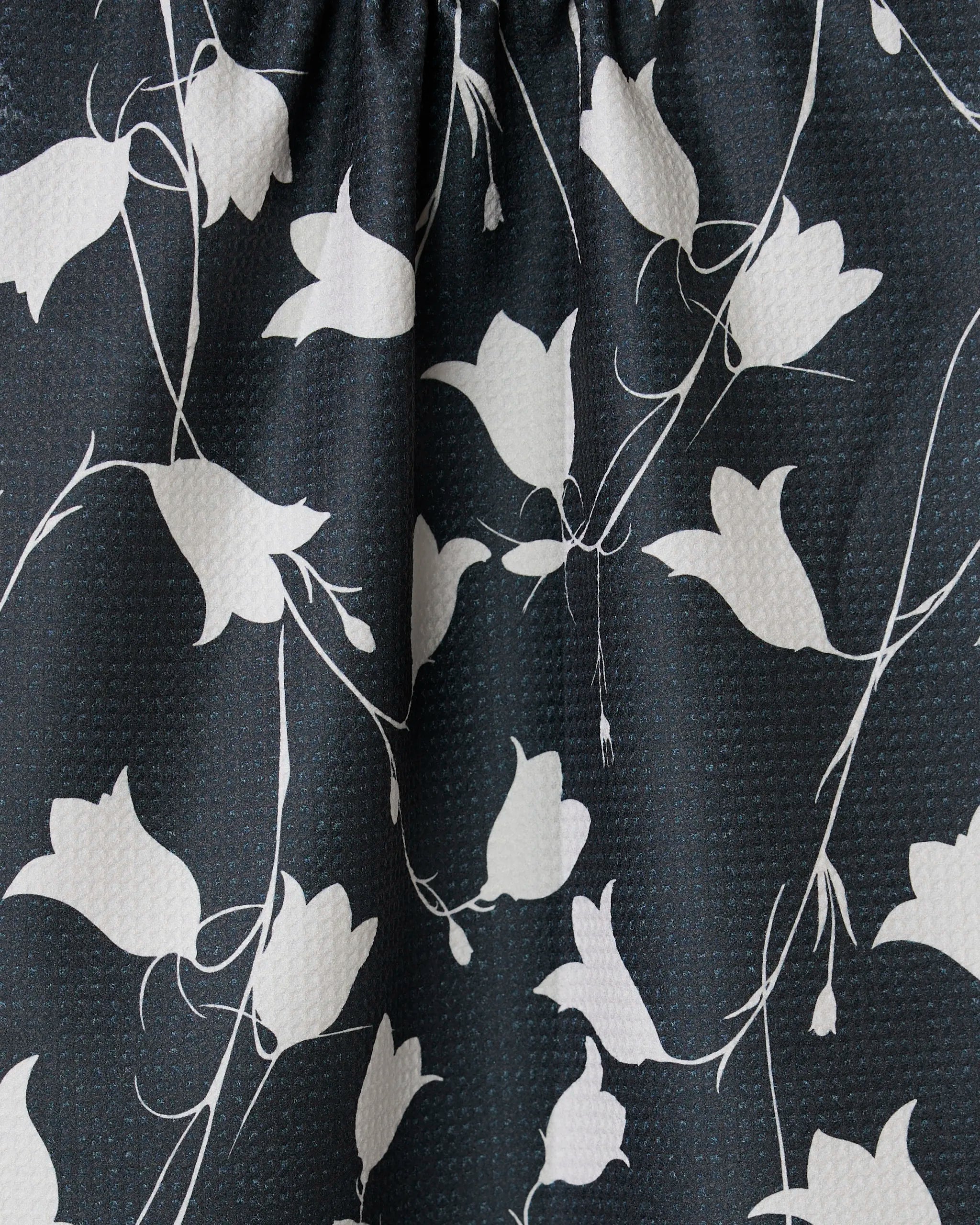 Close-up of GEOMETRYs Dancing Dark Flora tea towel, showcasing black fabric with a textured surface and a white pattern of abstract floral designs and slender vines.