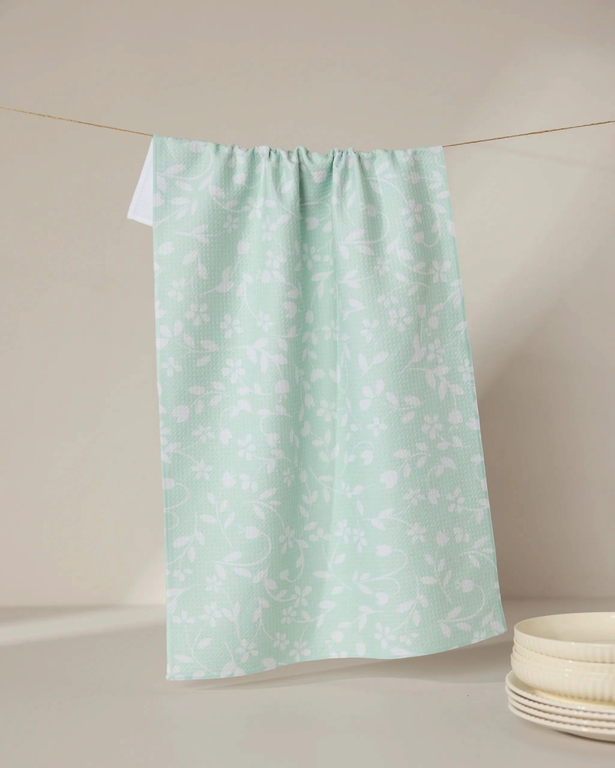 The GEOMETRY Dandy kitchen tea towel, light green with a white floral pattern and made from recycled materials, hangs on a clothesline. Ultra-absorbent and eco-friendly, it’s pictured with white bowls in the lower right corner.