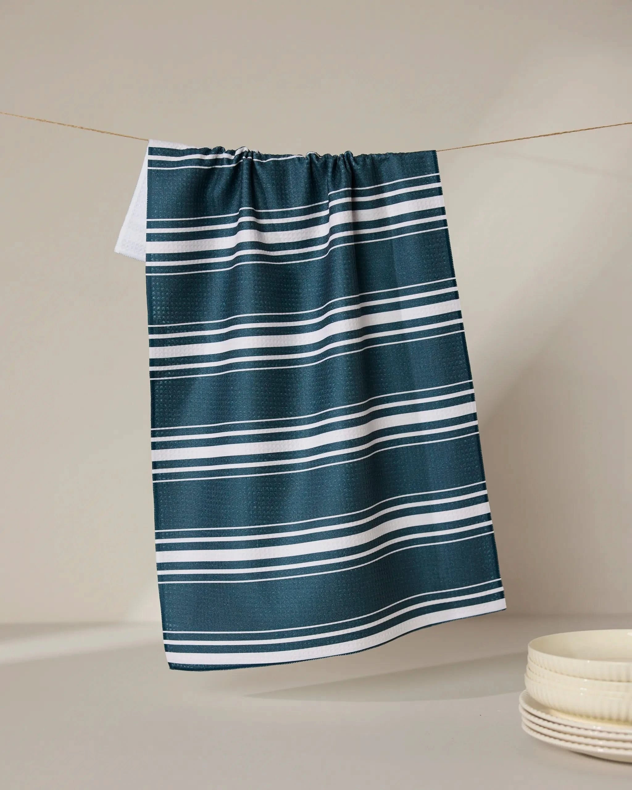 The GEOMETRY Deep Blue Fall recycled kitchen towel, featuring white horizontal stripes, hangs on a string against a neutral backdrop with stacked white ceramic dishes in the lower right corner.