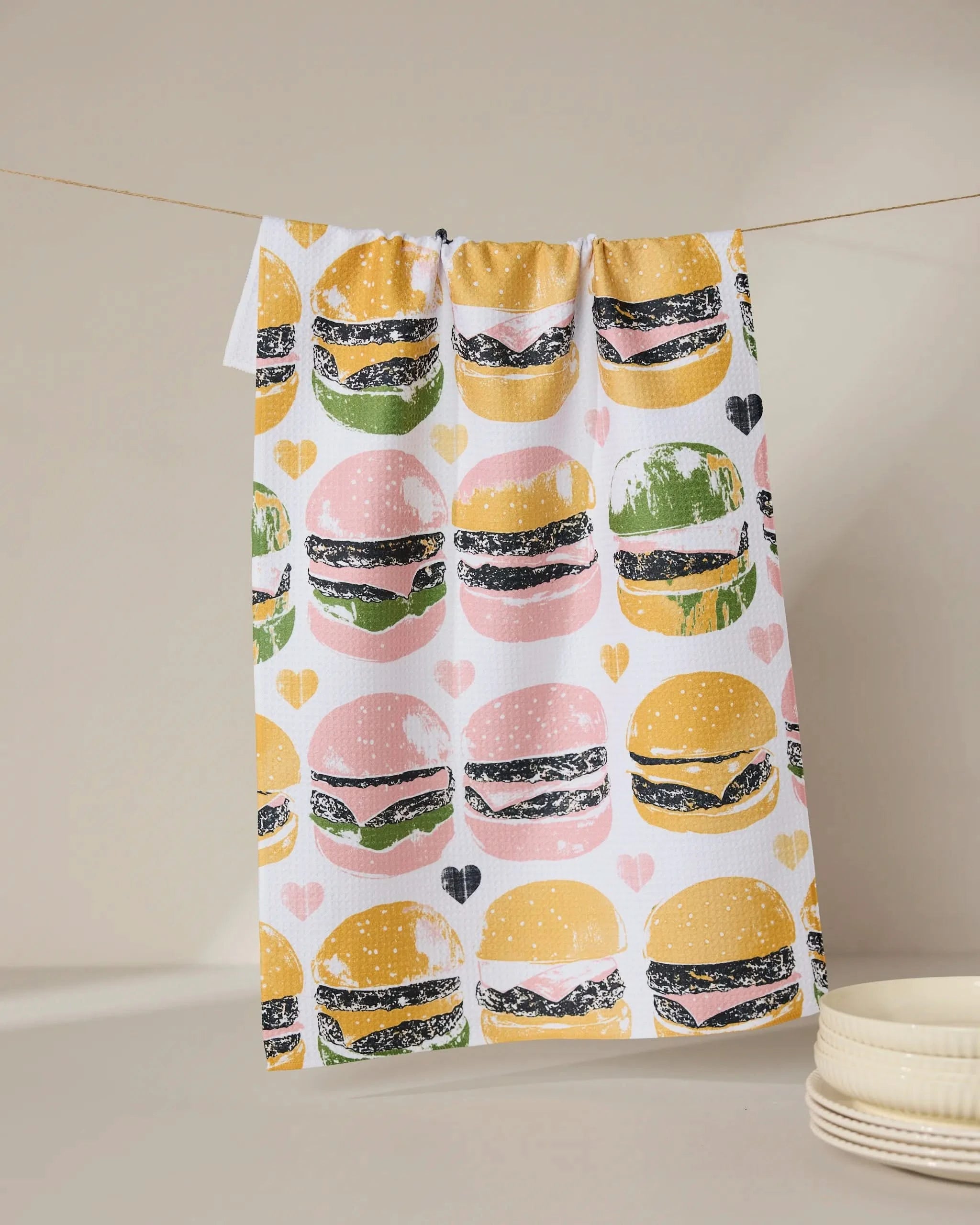 The Geometry On The Grill tea towel, featuring a vibrant hamburger and heart pattern, hangs above stacked white plates against a neutral backdrop.