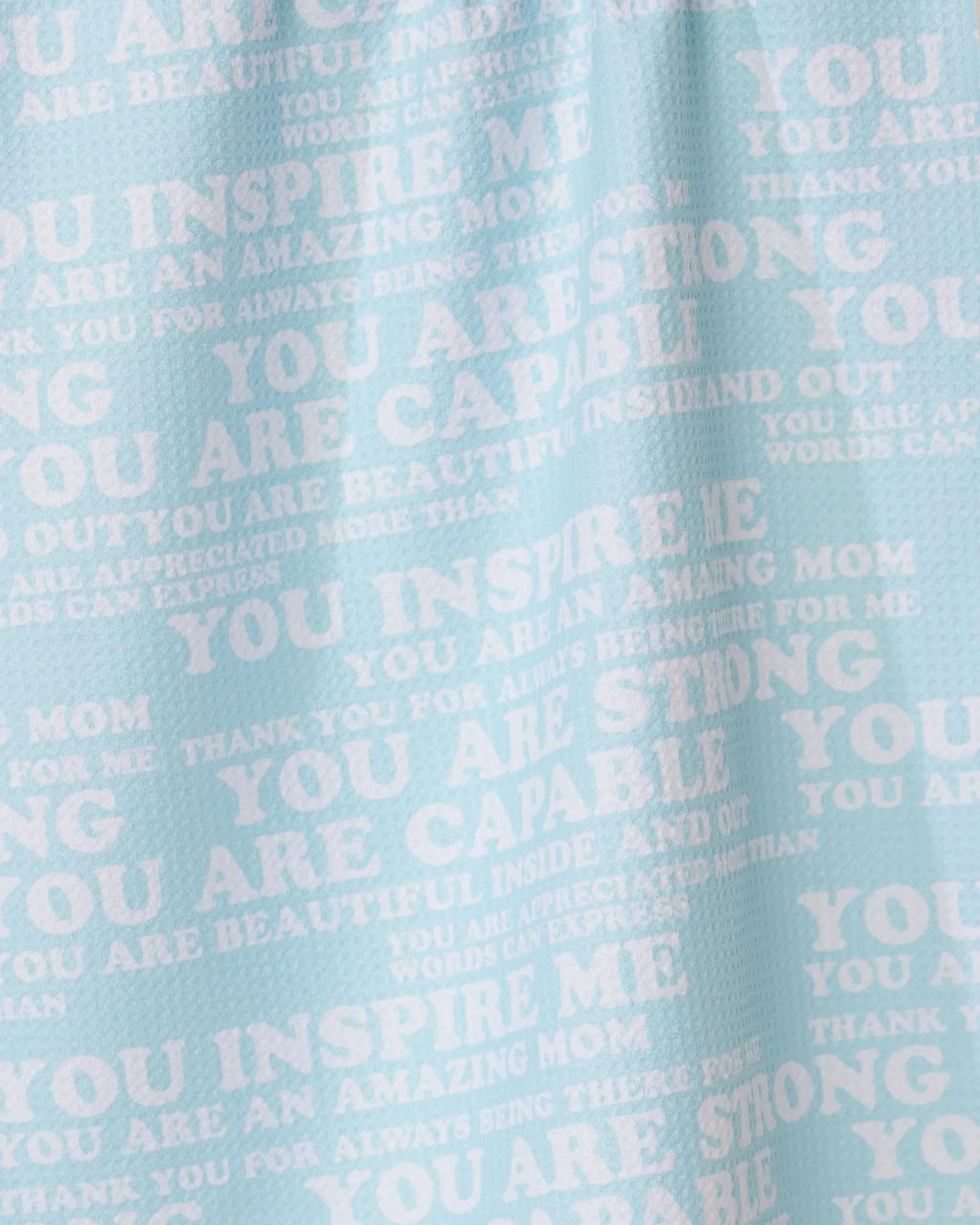 The GEOMETRY Mom Mantra tea towel is light blue, made from recycled materials, and features white overlapping motivational phrases like YOU INSPIRE ME and YOU ARE AMAZING MOM in different fonts and sizes.