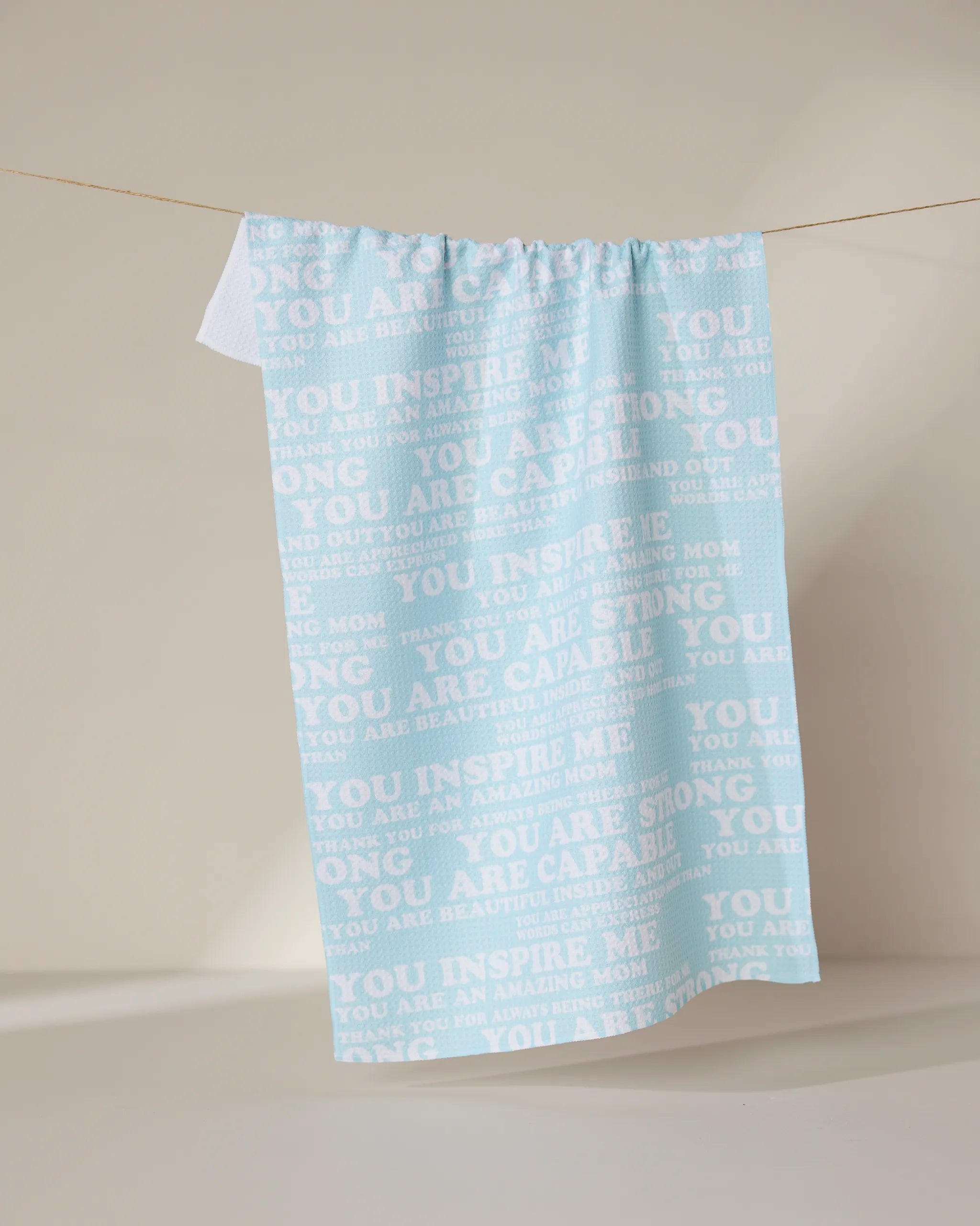 The GEOMETRY Mom Mantra is a light blue kitchen tea towel made from recycled materials, featuring white motivational phrases such as “You inspire me,” “You are strong,” and “You are capable.”.