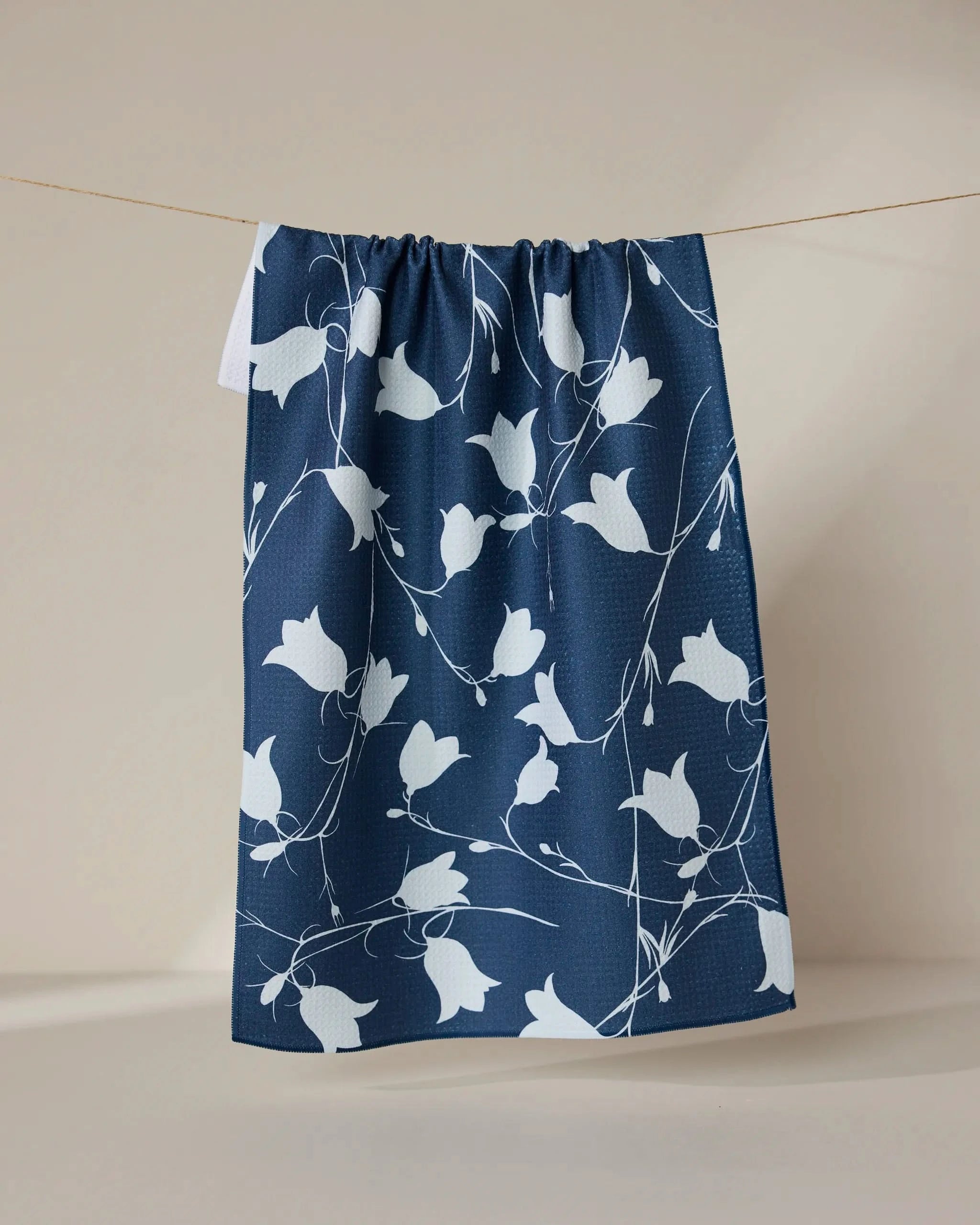 The GEOMETRY Navy Dancing Flora towel, featuring a white floral pattern on navy blue, hangs from a thin string against a plain beige background.