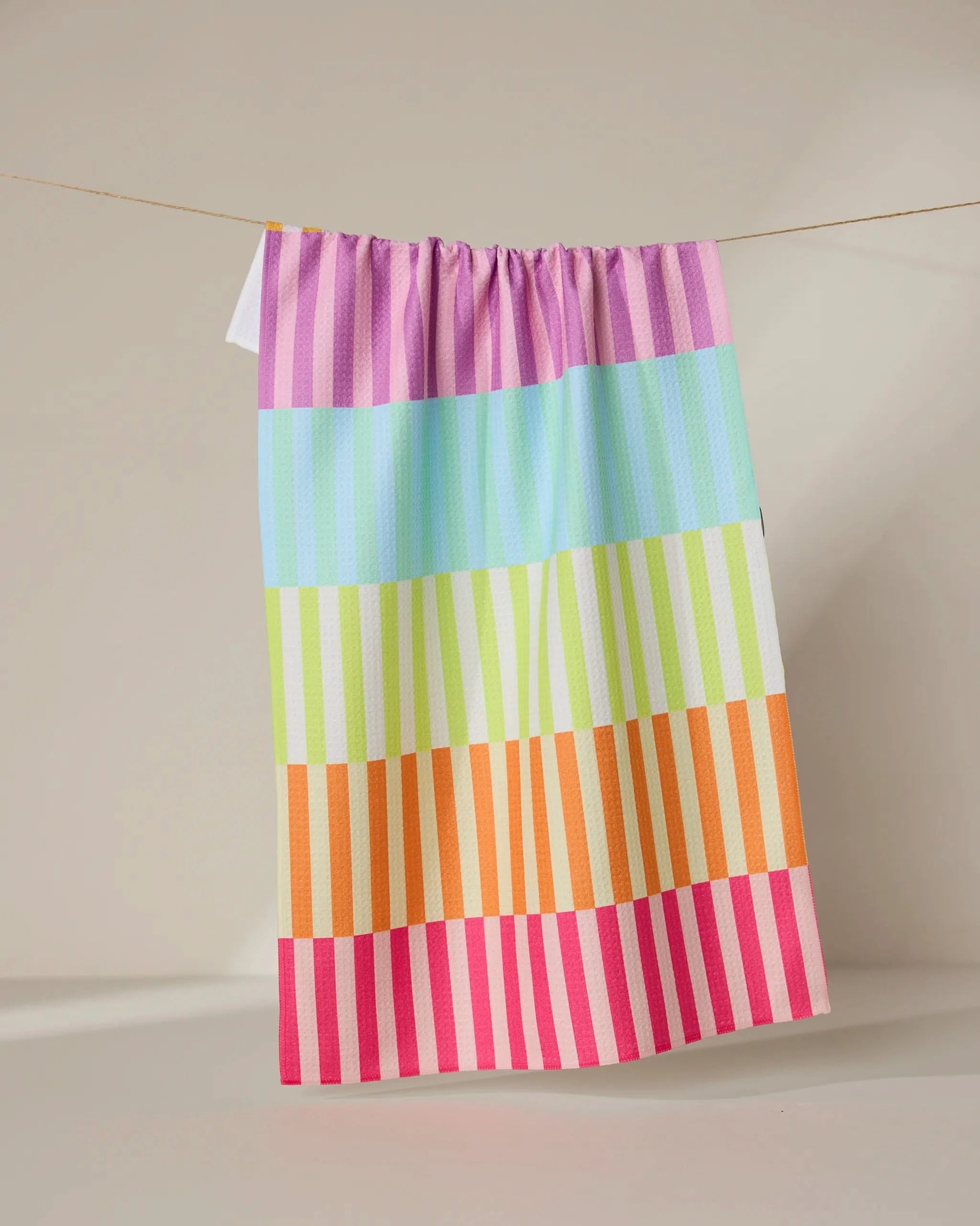 The GEOMETRY Neon Nights towel, featuring vibrant horizontal stripes in purple, blue, green, orange, and pink, hangs on a line against a softly lit neutral backdrop.