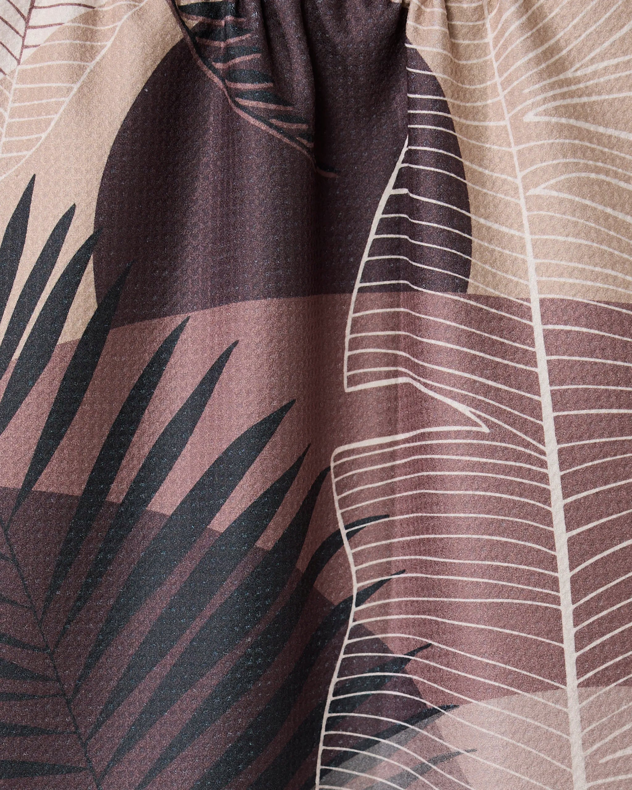 Close-up of the GEOMETRY Neutral Hills quick-drying towel, showcasing a printed pattern of overlapping abstract palm and fern leaves in beige, brown, and black with light gathering at the top.