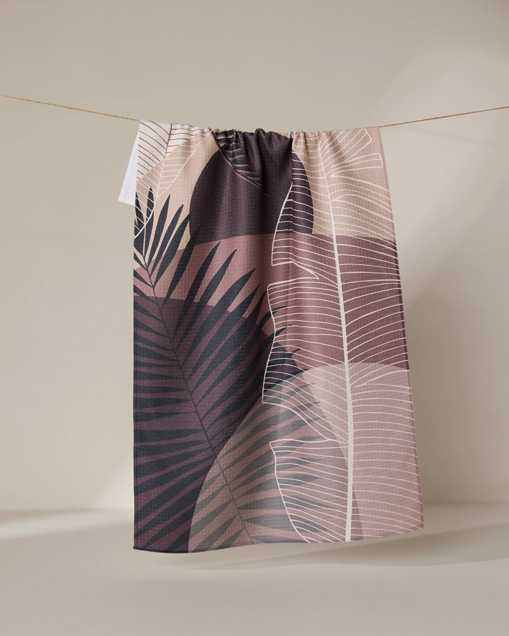 The GEOMETRY Neutral Hills tea towel, featuring a pink, mauve, and beige botanical leaf design, dries quickly and hangs on a string against a light beige background.