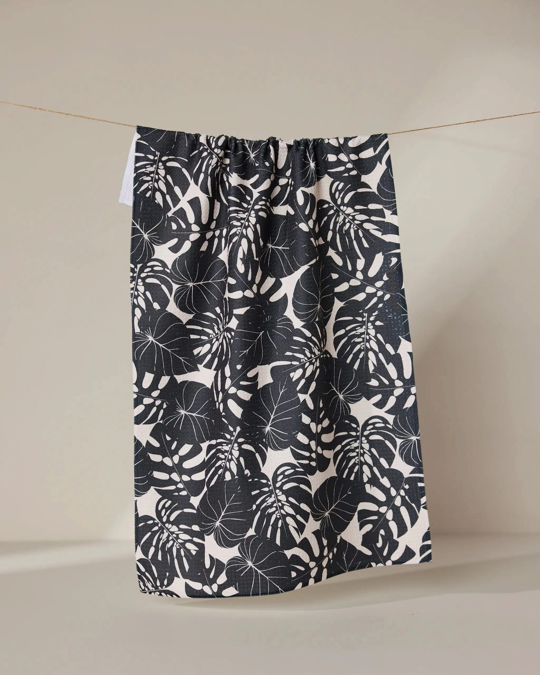 The GEOMETRY Neutral Leaves tea towel, featuring a bold black and white monstera leaf design and made from recycled materials, hangs on a clothesline against a plain beige background.