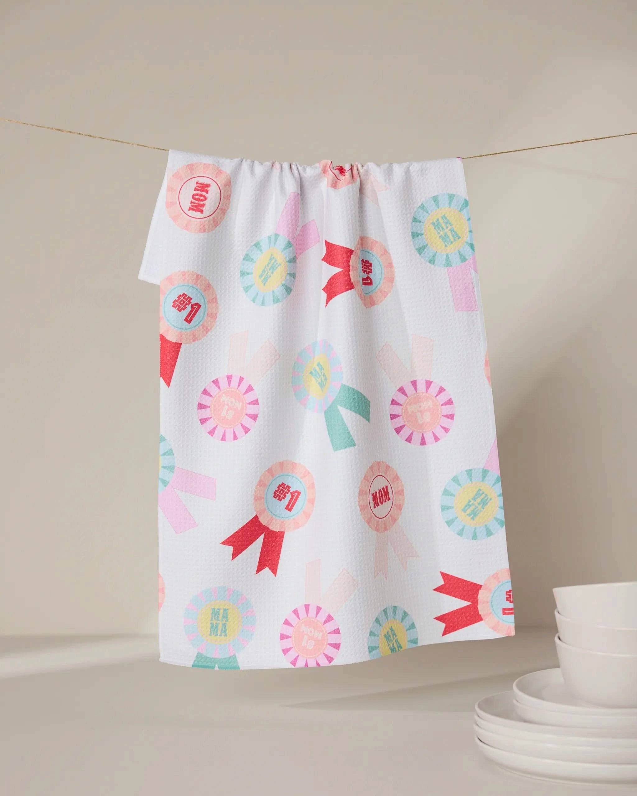 The “Number One Mom” tea towel by GEOMETRY, made from recycled materials, features colorful ribbon designs and “MOM” and “MAMA” text. It hangs above white dishes, offering an eco-friendly accent to a neutral kitchen.