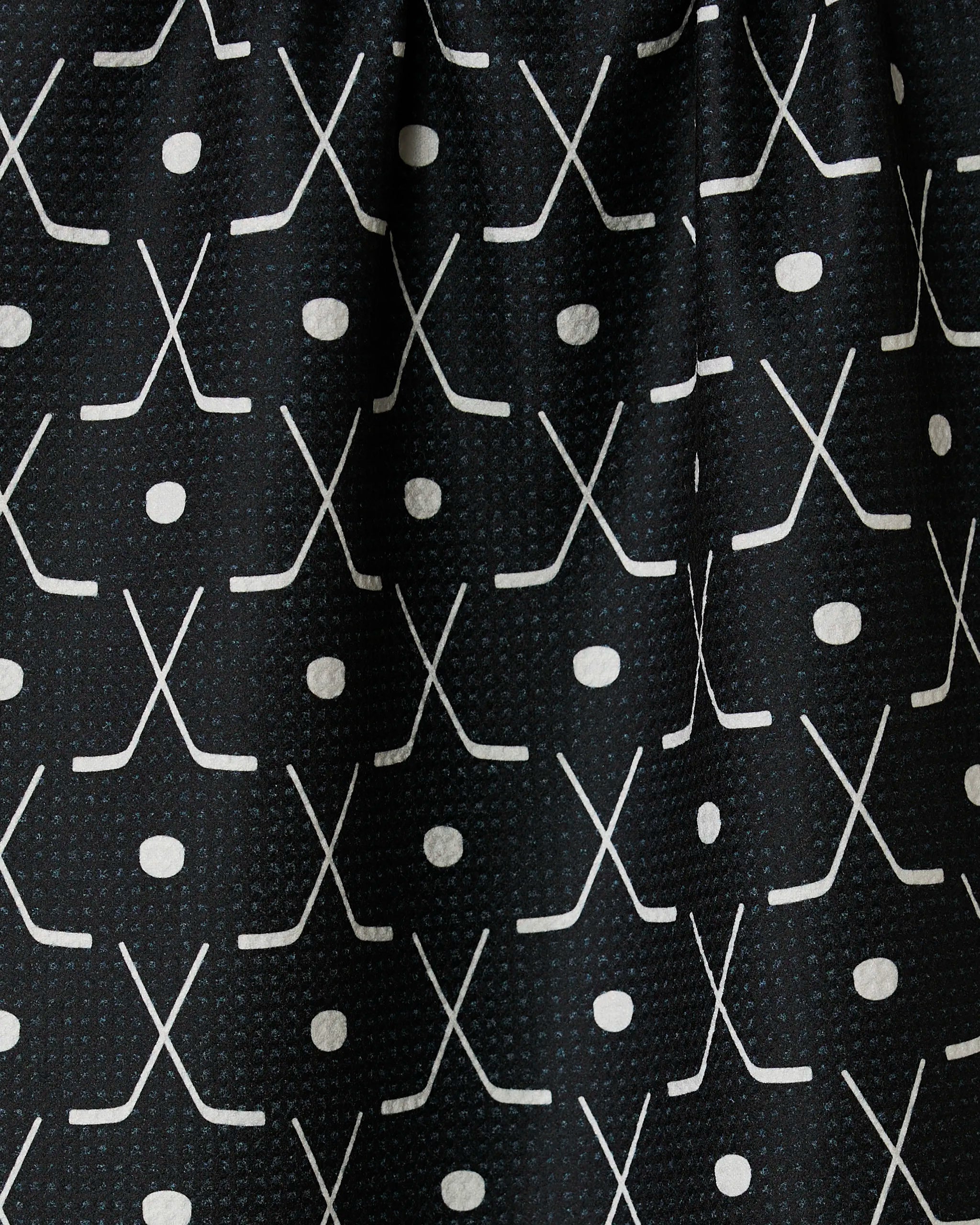 Close-up of the GEOMETRY Puck tea towel with ultra-absorbent black fabric, featuring a repeating grid pattern of white crossed hockey sticks and pucks for a sporty look.