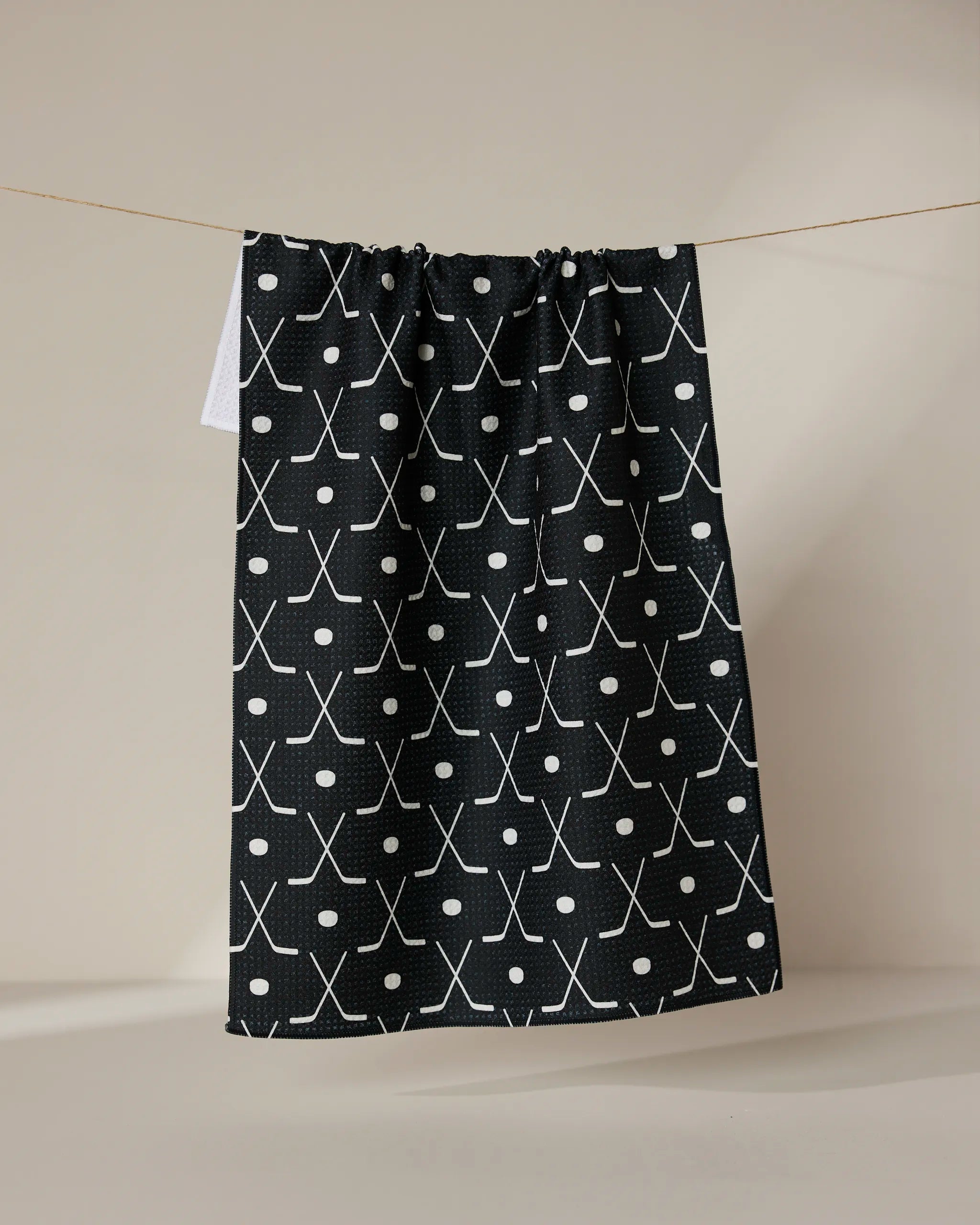 The GEOMETRY Puck tea towel is ultra-absorbent, showcasing a white crossed hockey stick and circle pattern on black, hanging on a string against a neutral background.