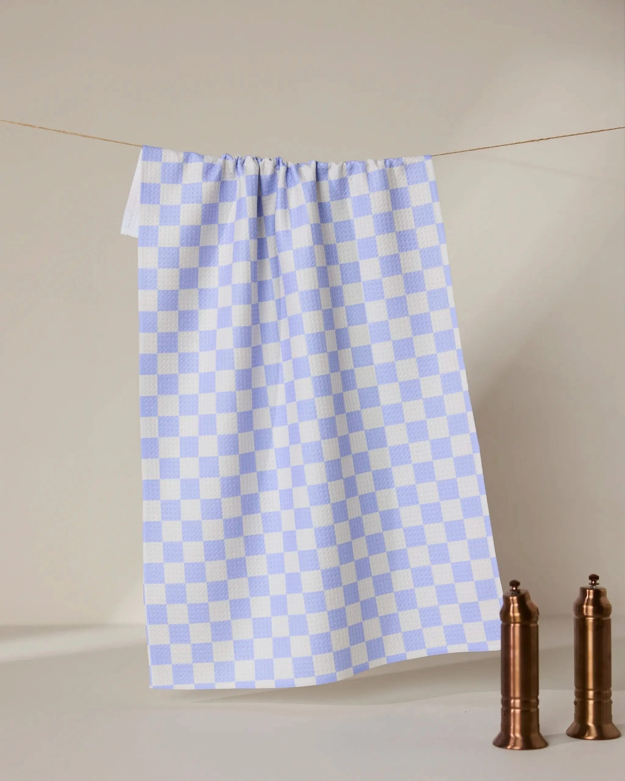 A Purple Picnic recycled kitchen towel by GEOMETRY hangs on a string against a beige background, with two brown mills on a white surface in the lower right corner.