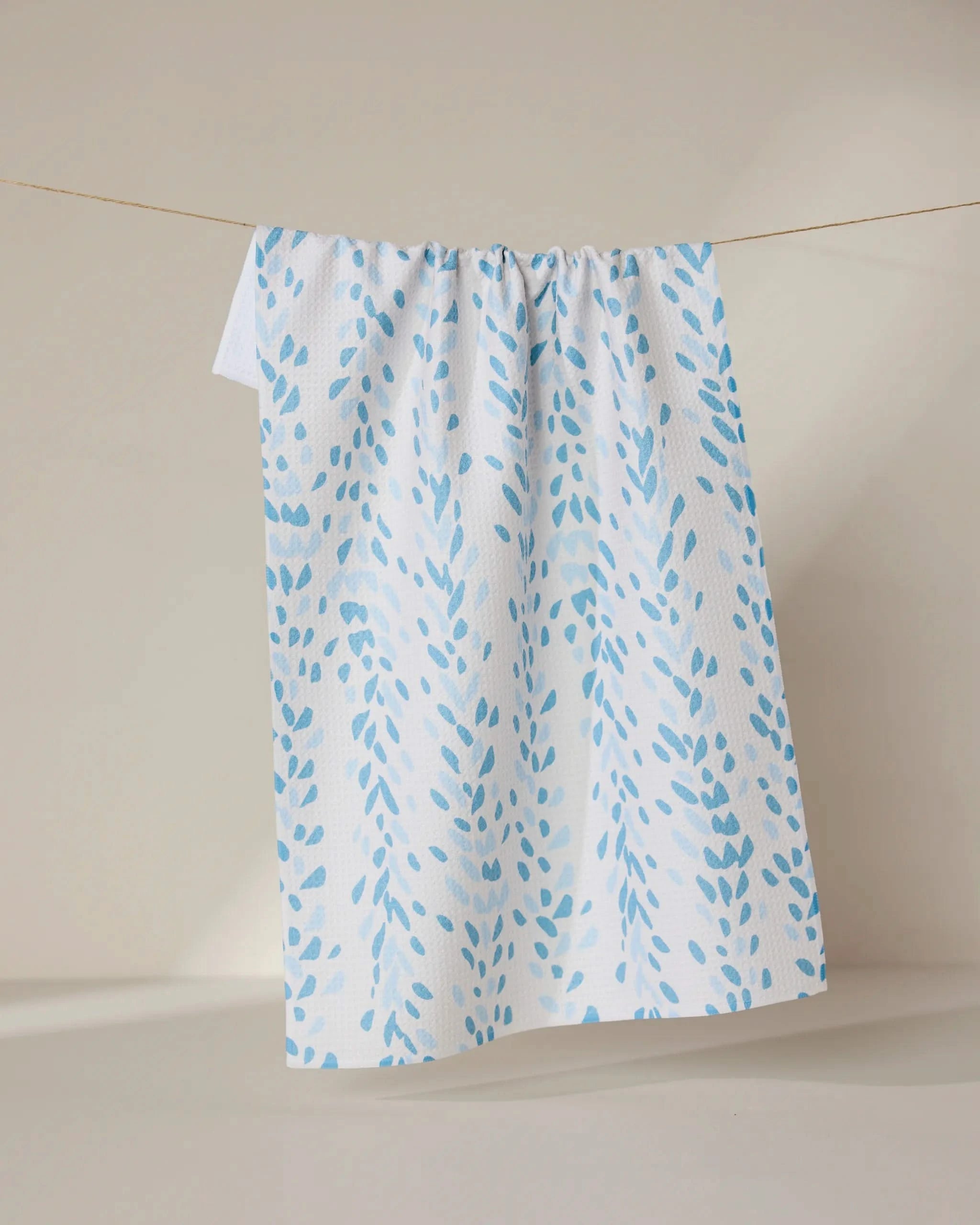 The GEOMETRY Reeds Printed - Midday is a white tea towel featuring light blue leaf-like patterns, crafted from recycled materials, shown hanging neatly on a clothesline against a neutral background.