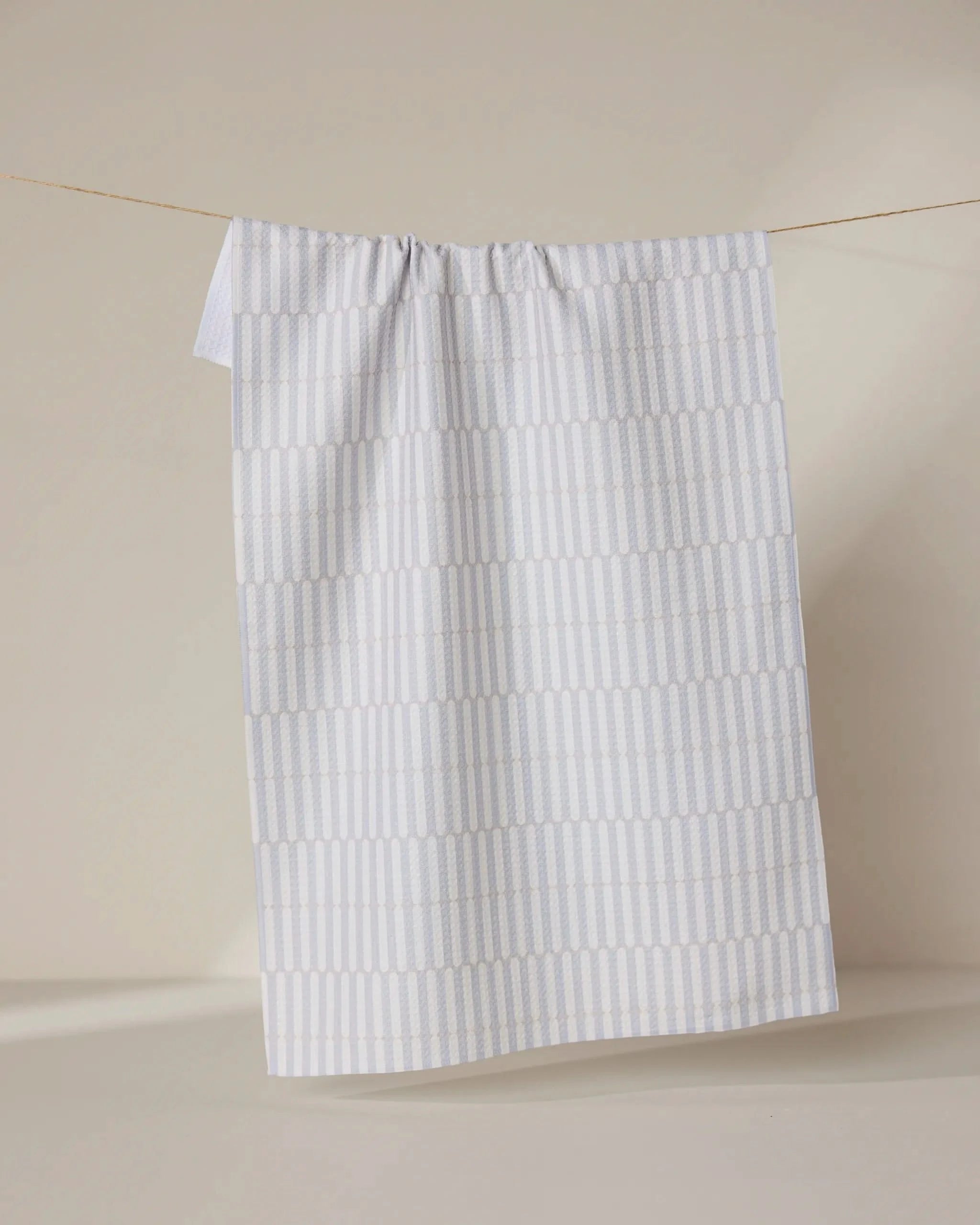 The GEOMETRY Rideaux Neutre tea towel, featuring a subtle light grey geometric pattern on white, is draped over a string against a neutral beige background.